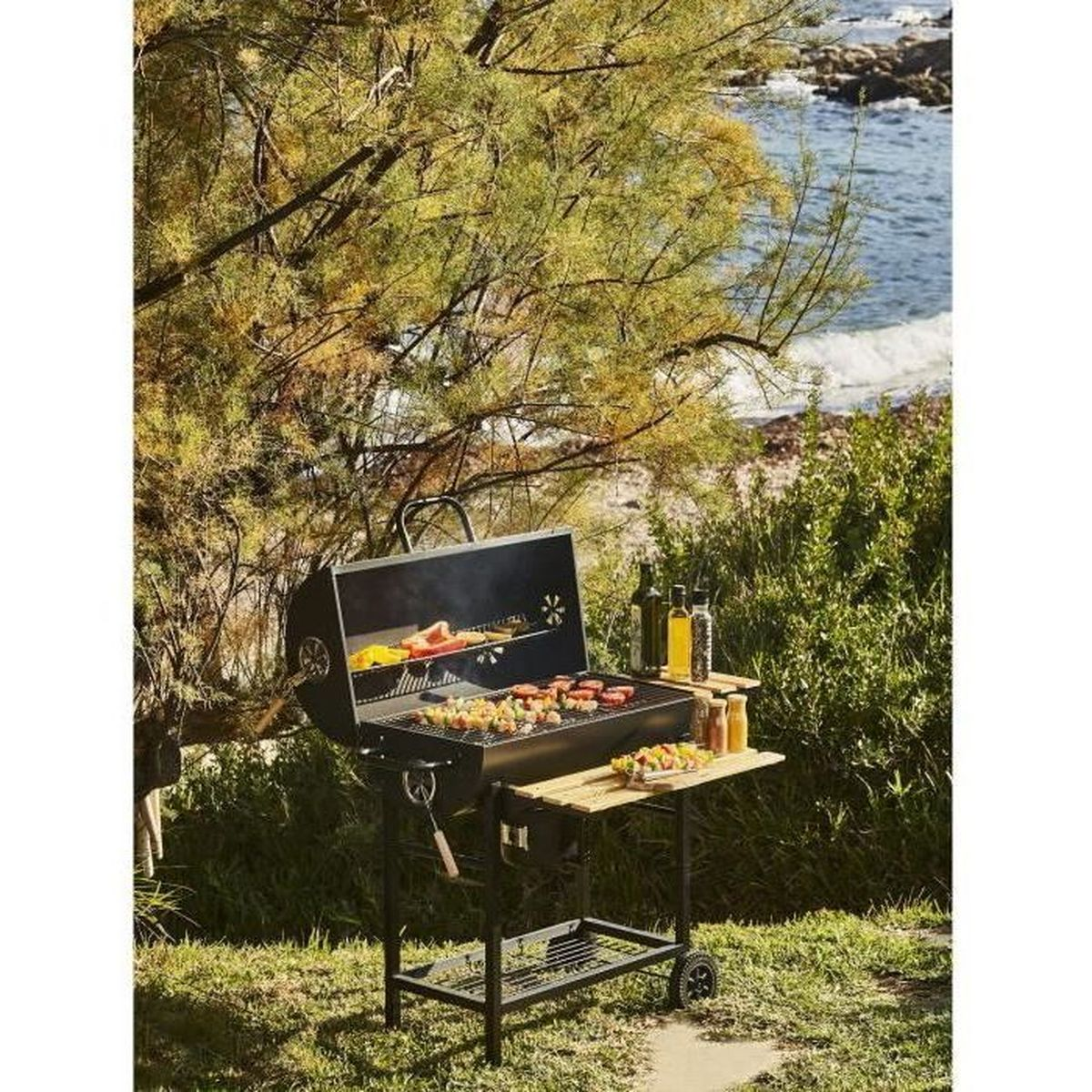 Een grill met eten aan zee onder een boom, met flessen en kruiden.