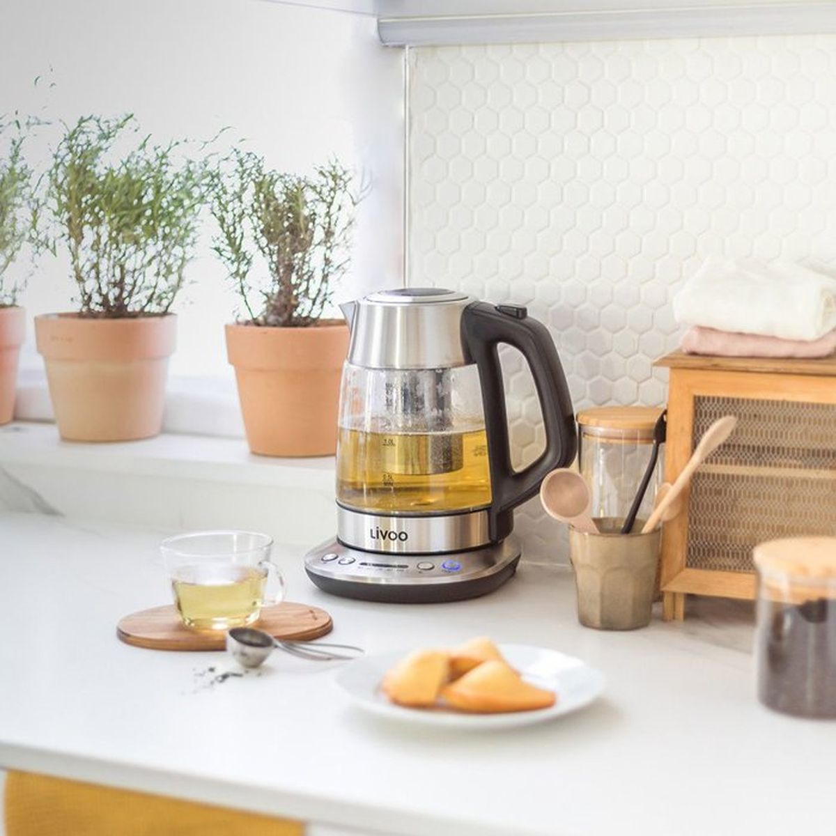 Un hervidor de cristal con té, taza, galletas y plantas en una encimera.