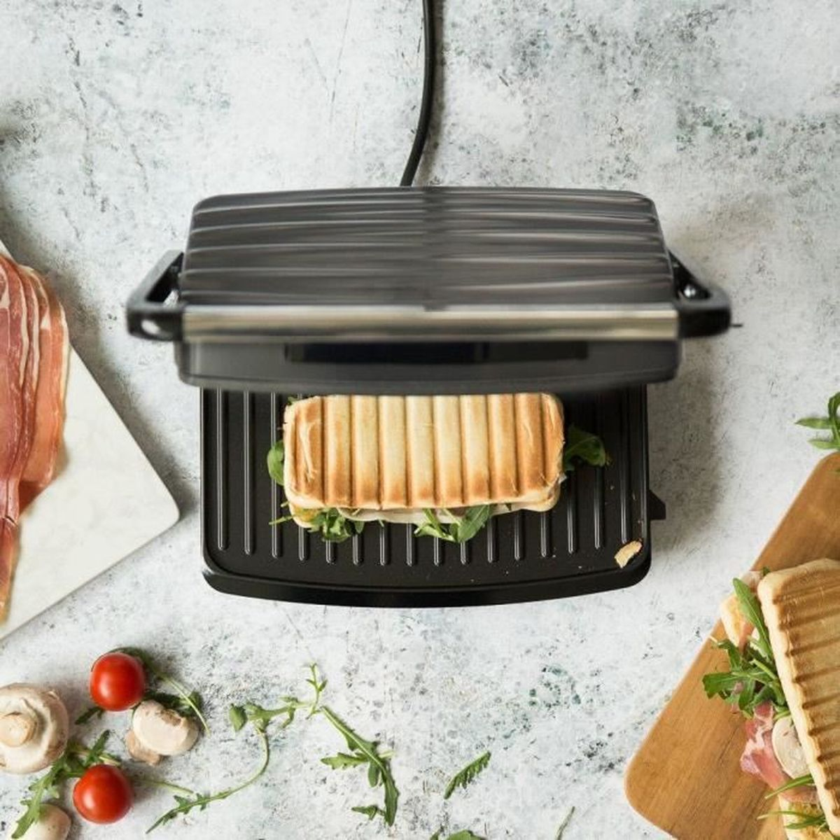 Un panino viene grigliato in una piastra per panini, con prosciutto, pomodori e funghi nelle vicinanze.