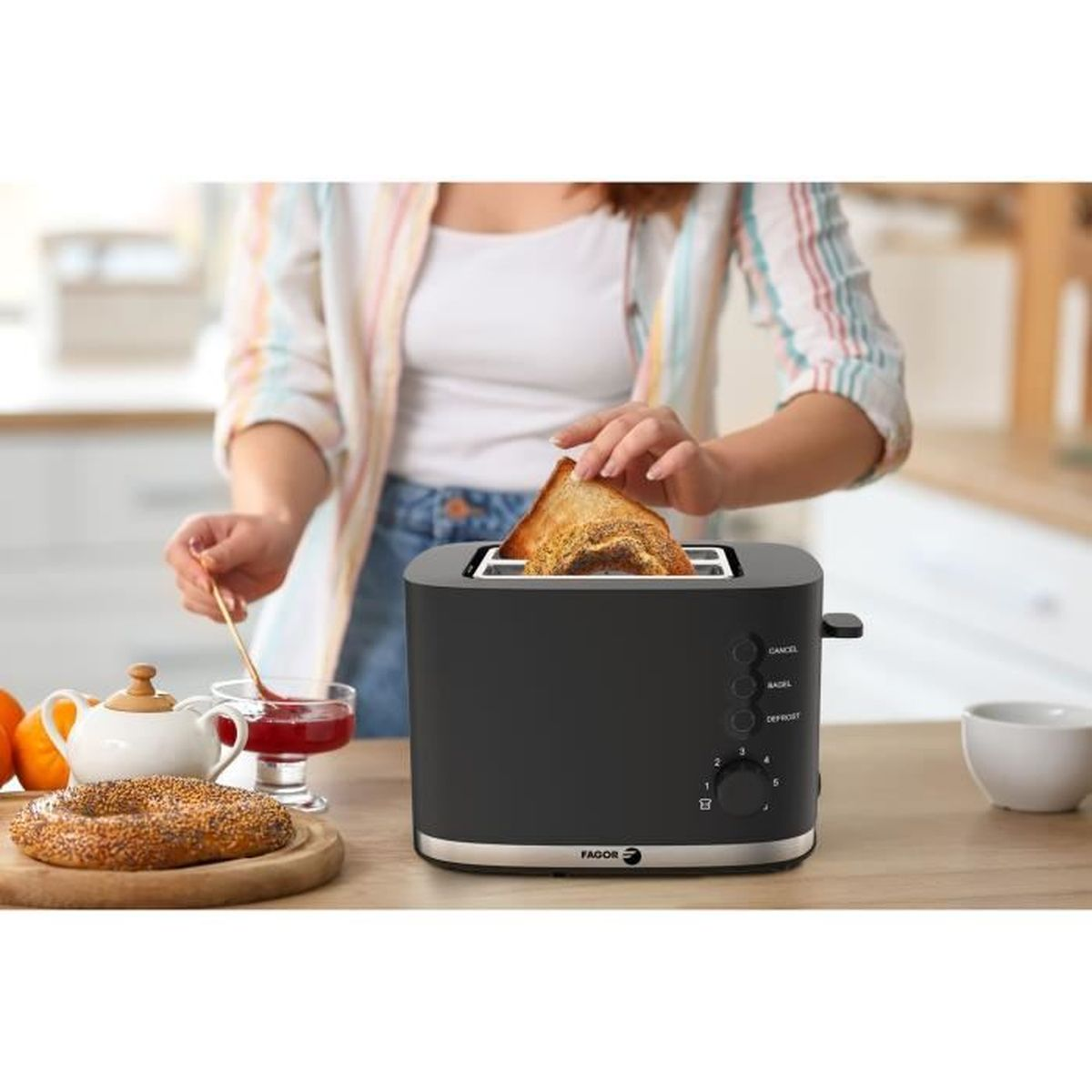 Eine Person nimmt Toast aus einem schwarzen Toaster, daneben ein Bagel und Marmelade.
