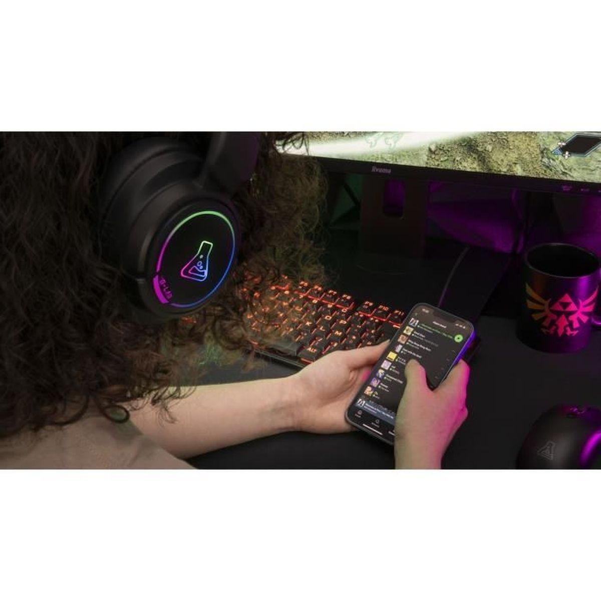 Osoba w słuchawkach trzyma smartfon, klawiatura i monitor w tle.