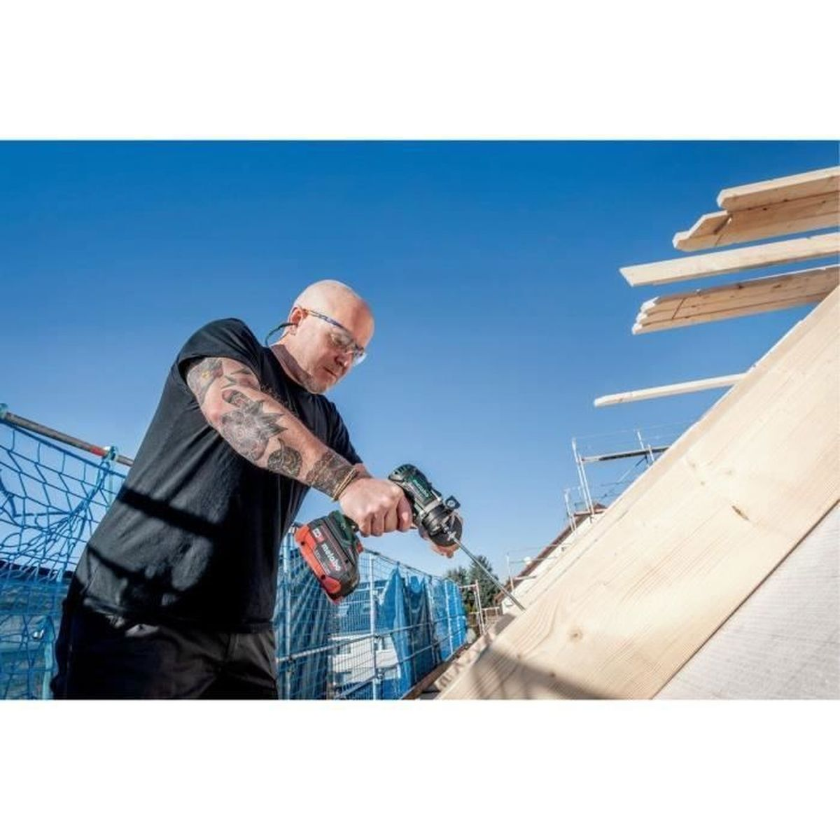 Mężczyzna z tatuażami wierci drewno na budowie pod błękitnym niebem.