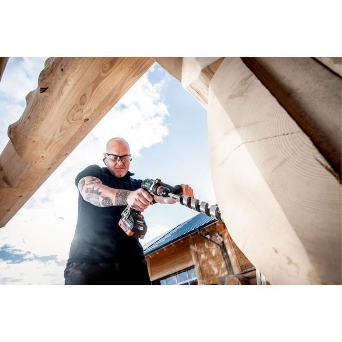 Mężczyzna wierci w drewnie dużym wiertłem. Błękitne niebo i budynek w tle.