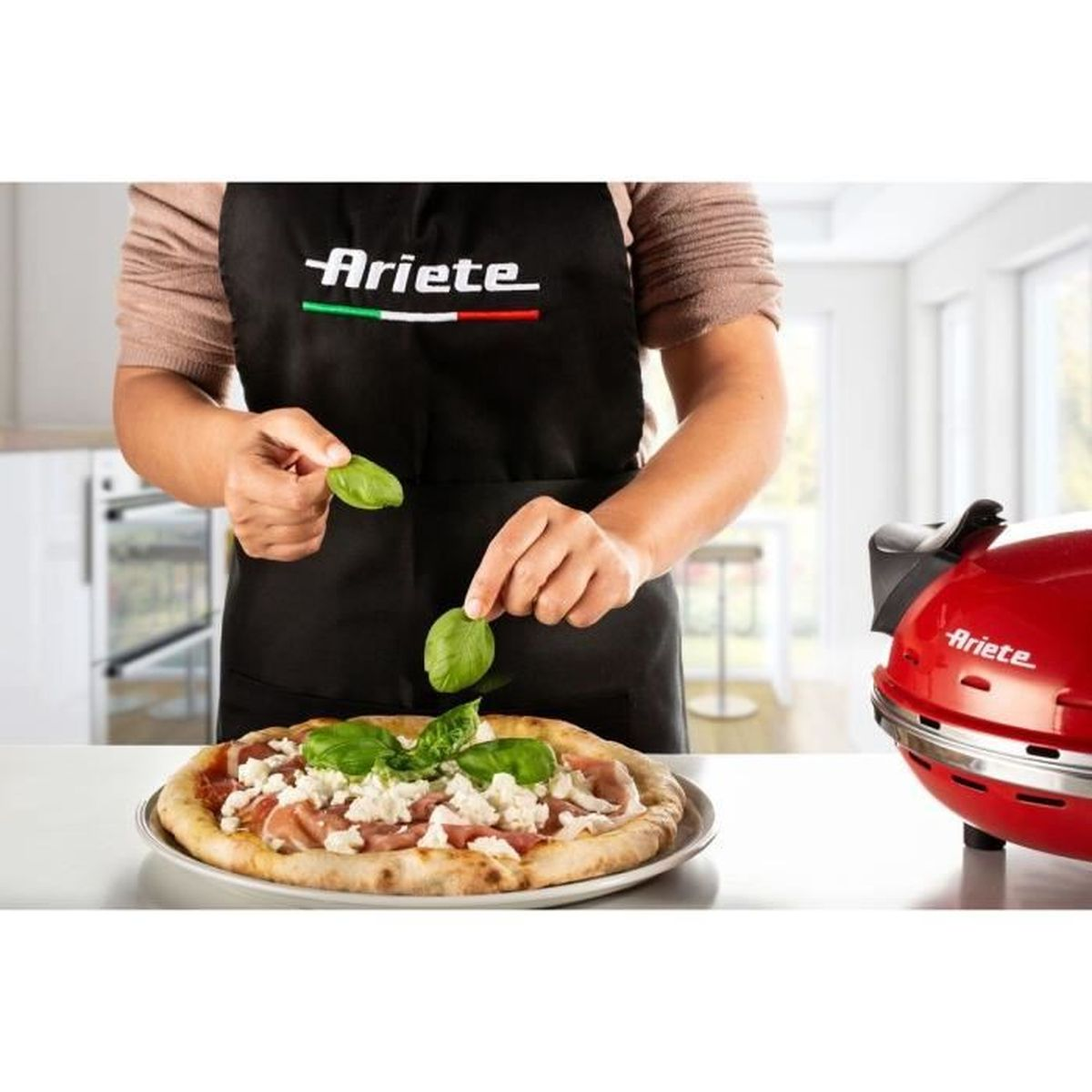 Een persoon garneert een pizza met basilicumblaadjes voor een rode pizzaoven.