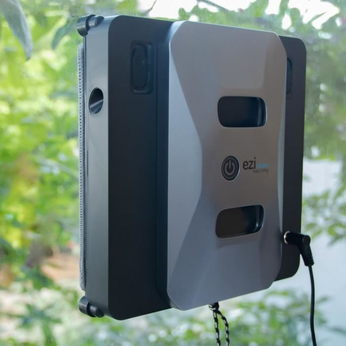 Robot do czyszczenia okien przylega do okna, z zielonymi liśćmi w tle.