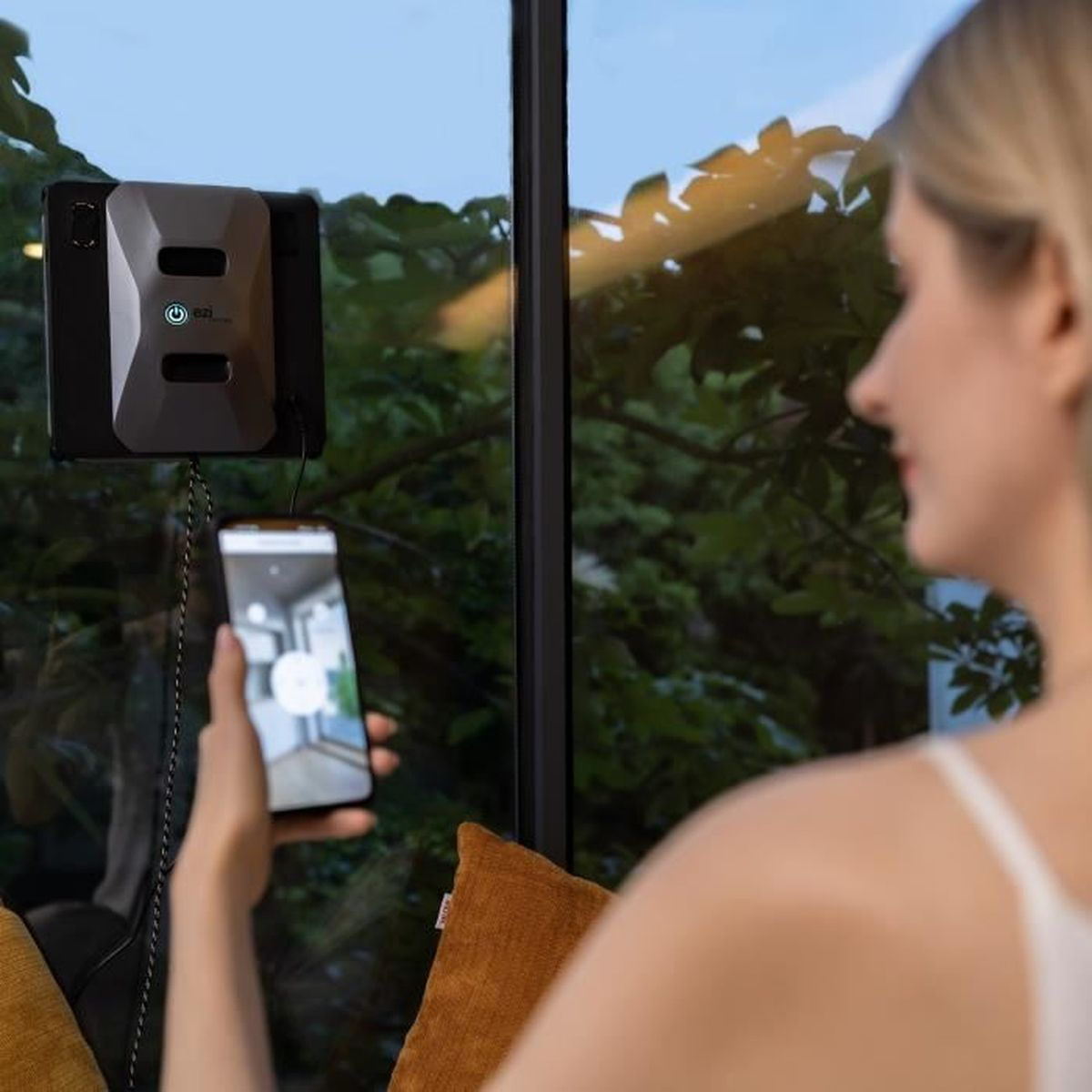 Robot do czyszczenia okien przylega do okna, podczas gdy kobieta steruje nim za pomocą smartfona.
