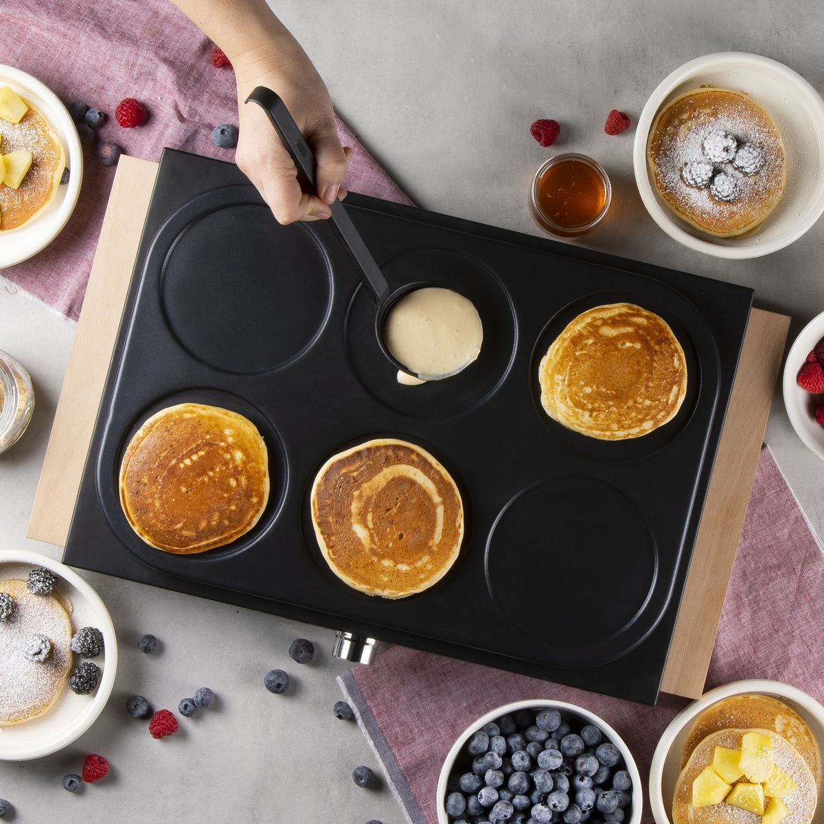 Eine Hand gießt Teig auf eine Pfannkuchenmaschine. Fertige Pfannkuchen, Beeren und Honig.