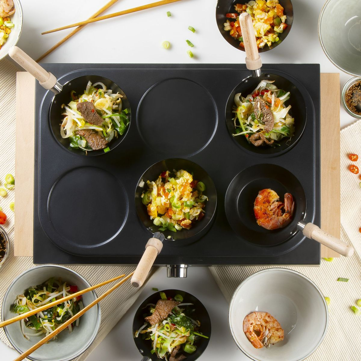 Mehrere kleine Pfannen mit asiatischem Essen auf einem Kochfeld, garniert mit Frühlingszwiebeln.