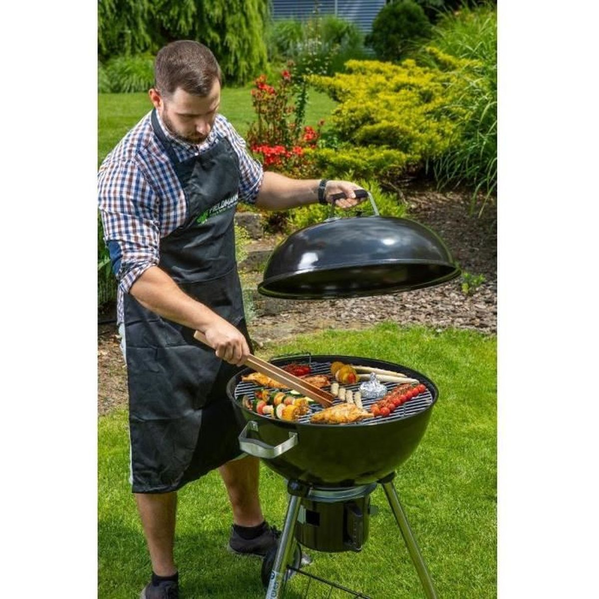 A man wearing an apron grills food on a round barbecue in a garden.
