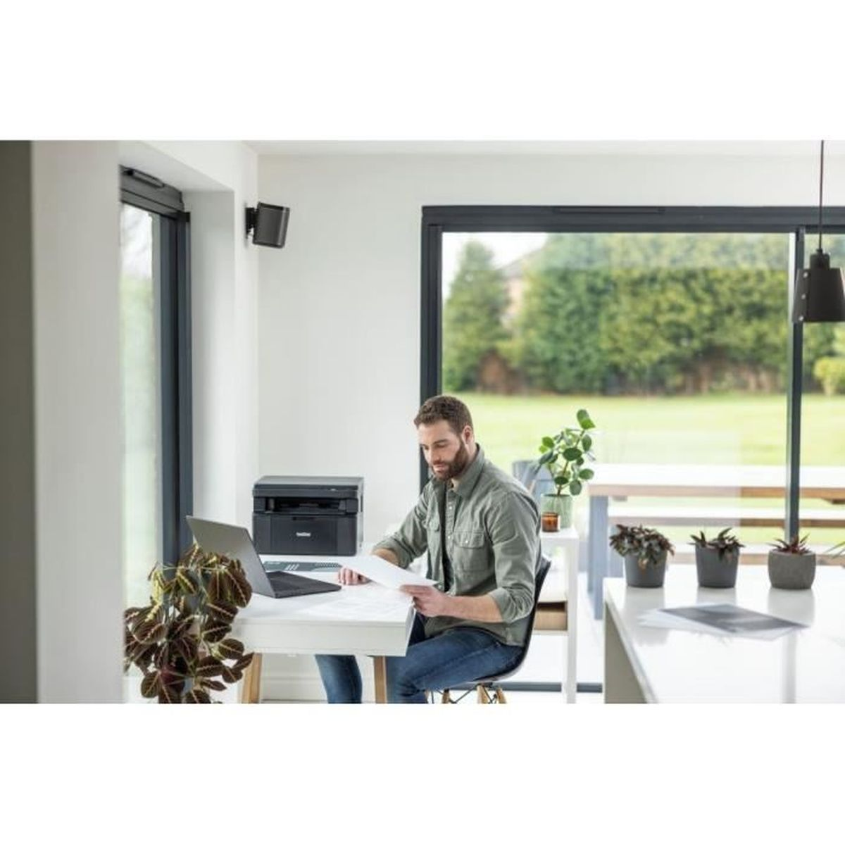 Een man zit aan een bureau, werkend aan documenten met een laptop en printer.
