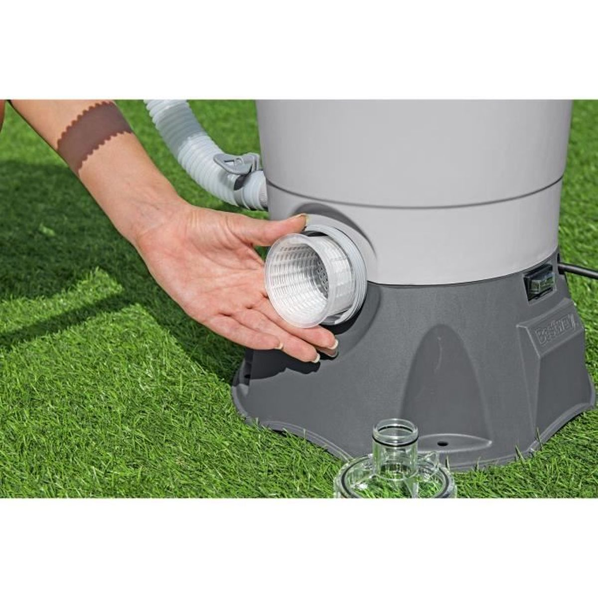 Ręka trzyma biały kosz filtra do szarej obudowy filtra basenowego na trawie.