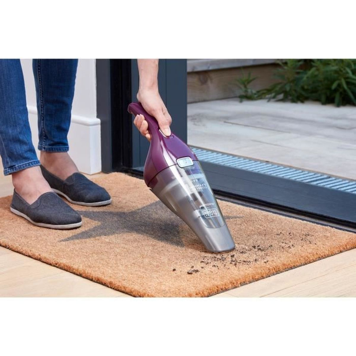 Odkurzacz ręczny czyści wycieraczkę. Widać niebieskie jeansy, szare buty.