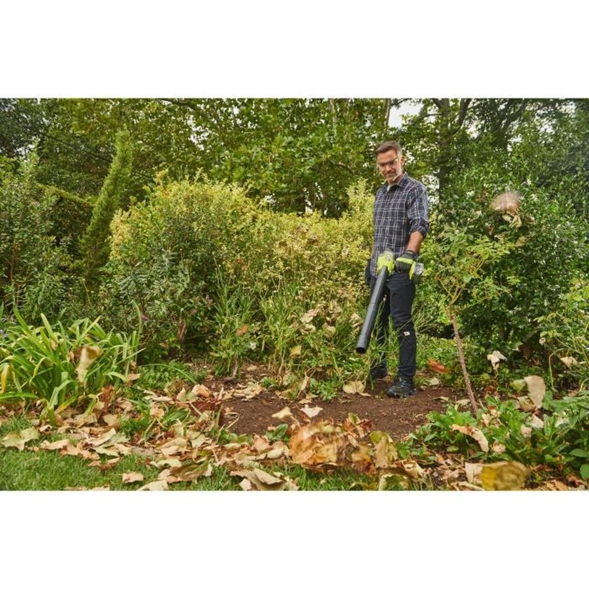Mann mit Laubbläser im Garten, umgeben von Büschen und gefallenen Blättern.