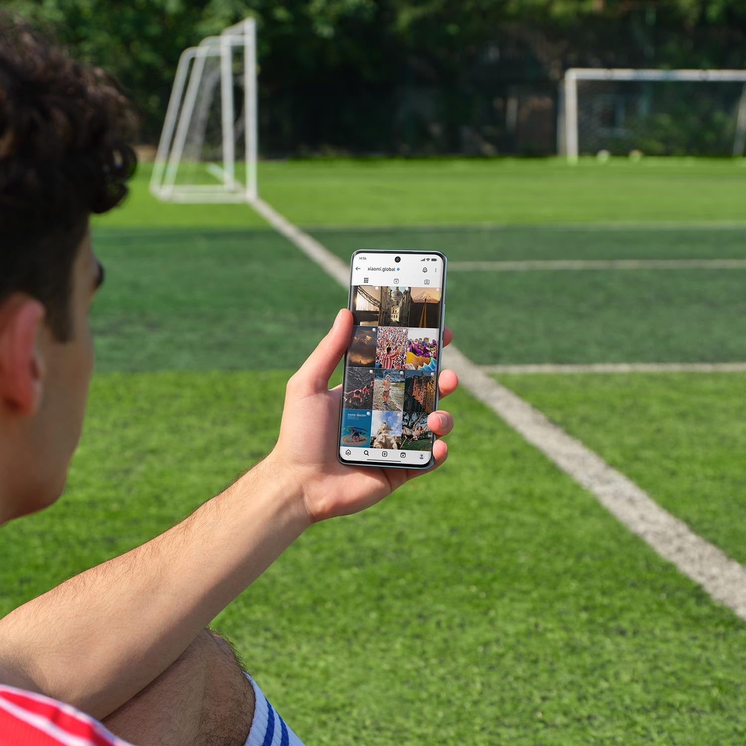 Ein Mann hält ein Smartphone mit Social-Media-Fotos vor einem Fußballplatz.