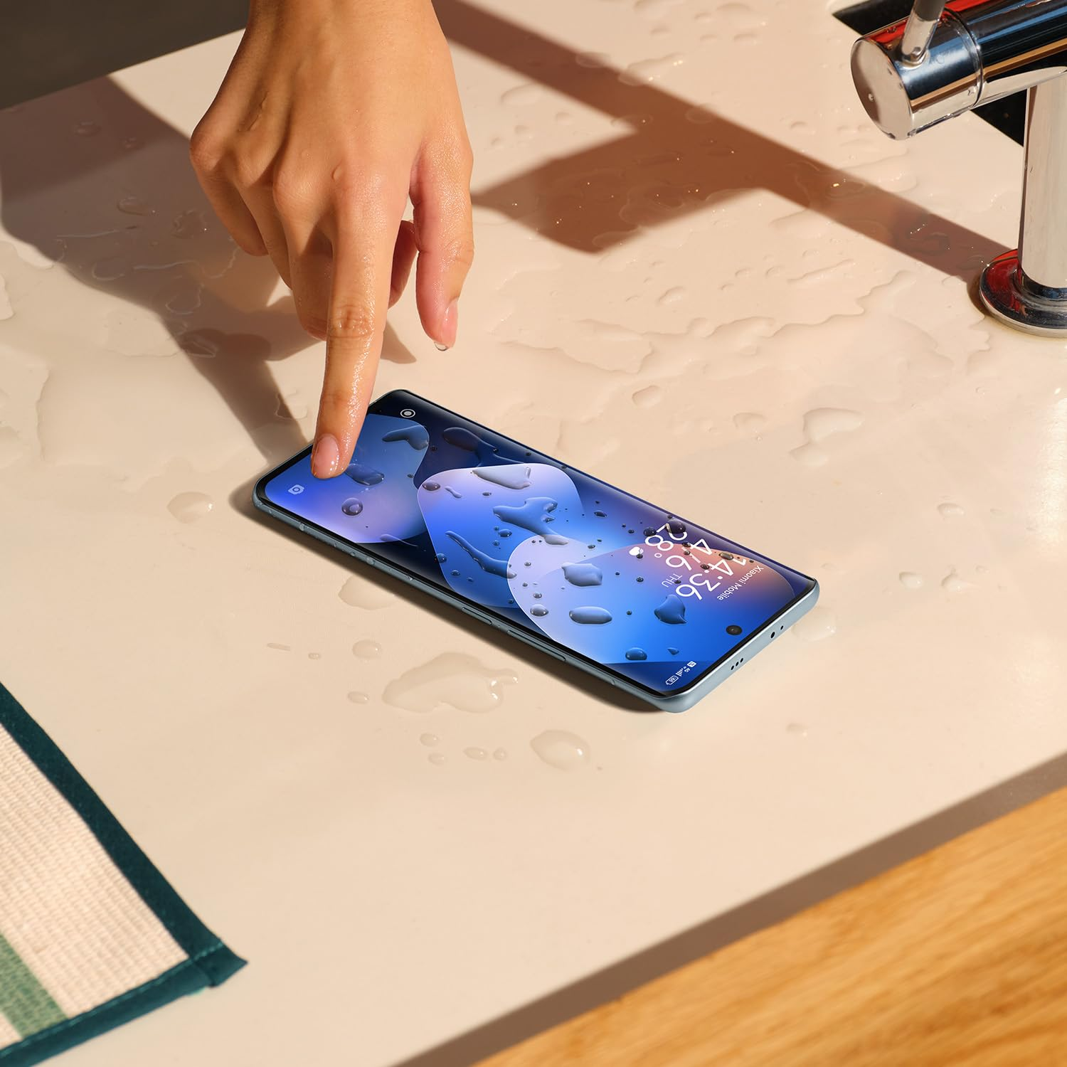 Eine Hand berührt ein nasses Smartphone auf einer hellen Oberfläche neben einem Wasserhahn.