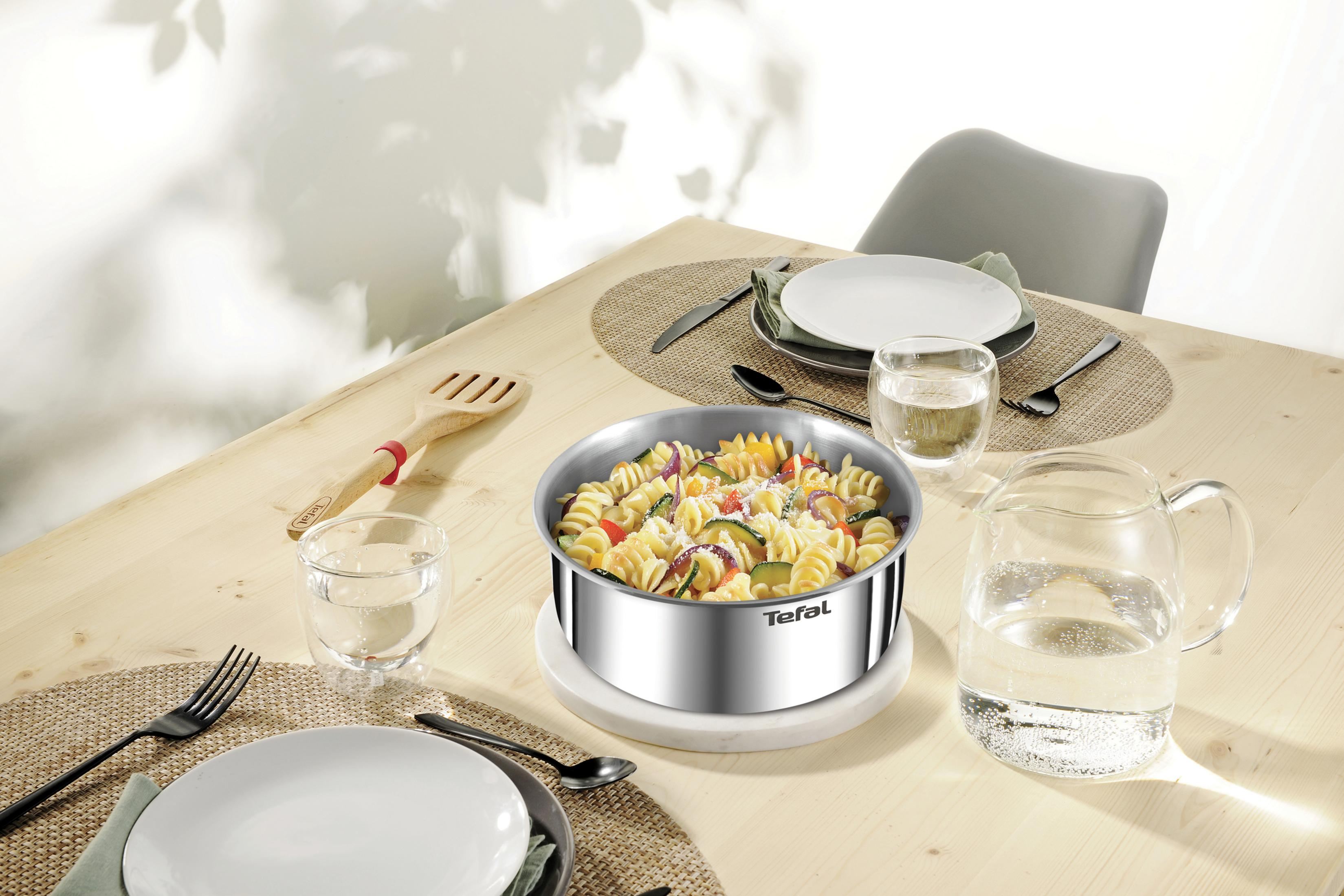 Una olla de pasta y verduras sobre una mesa de madera con vajilla.