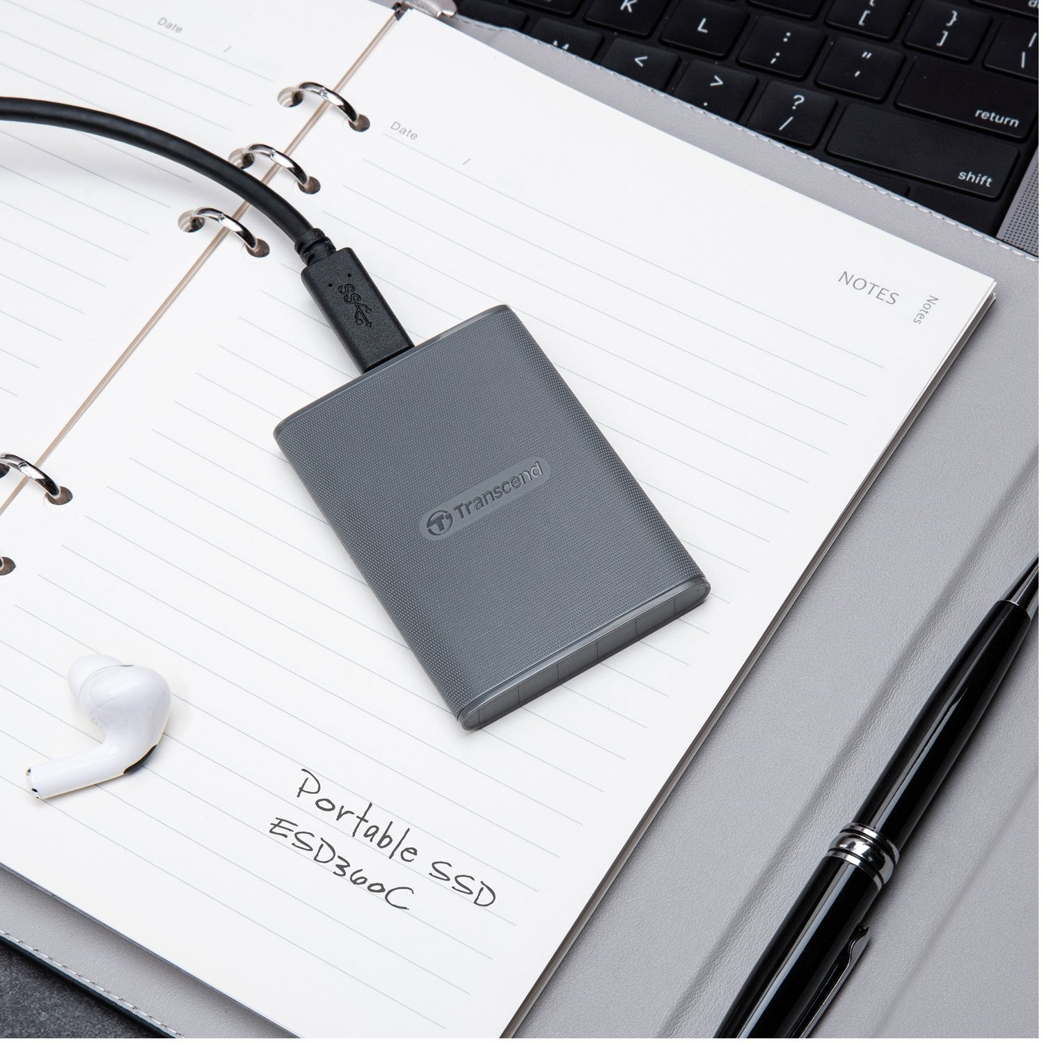 SSD portatile grigio collegato via cavo USB a un taccuino, con auricolari e penna.