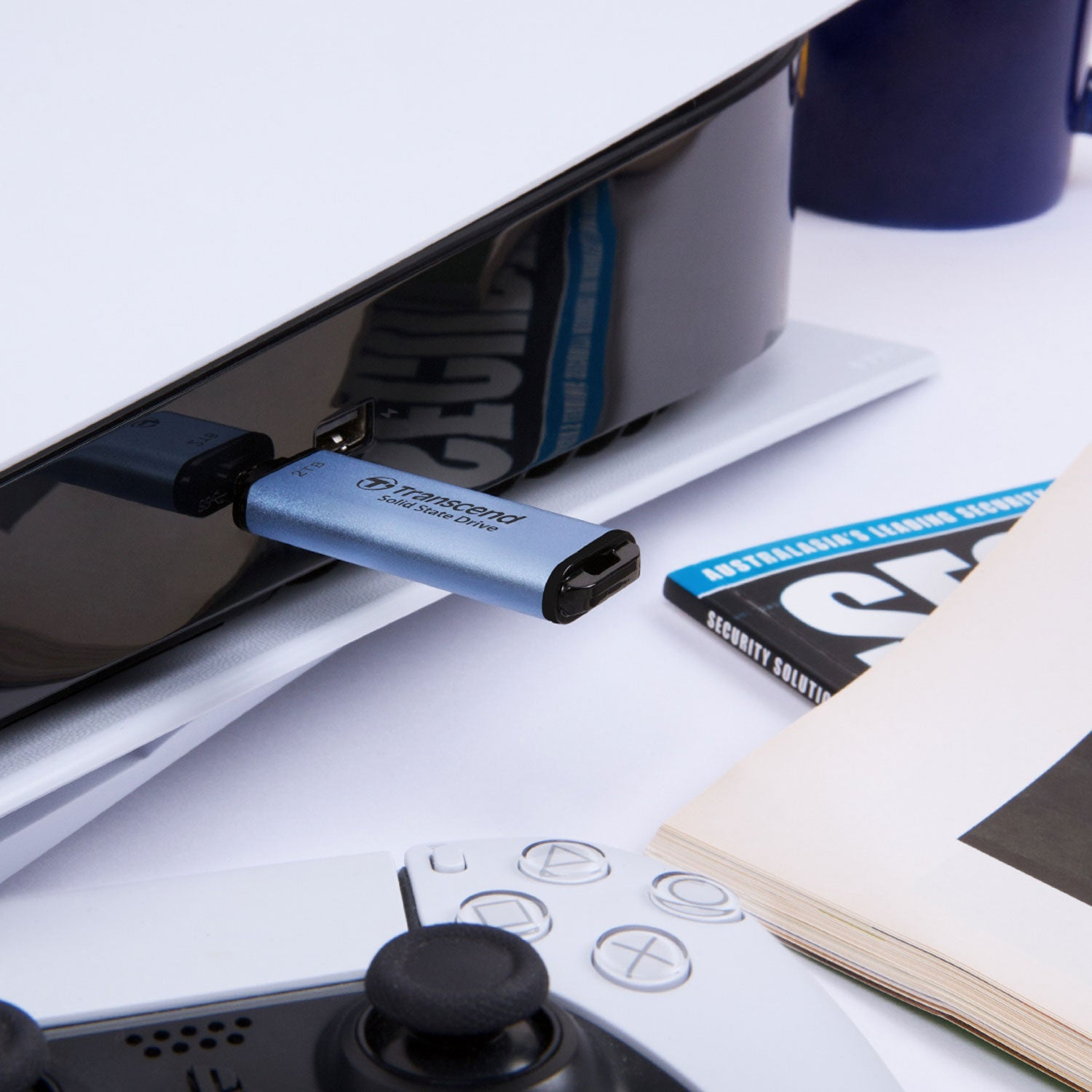 Niebieski pendrive Transcend jest podłączony do czarnej konsoli do gier. W pobliżu znajduje się kontroler i książka.