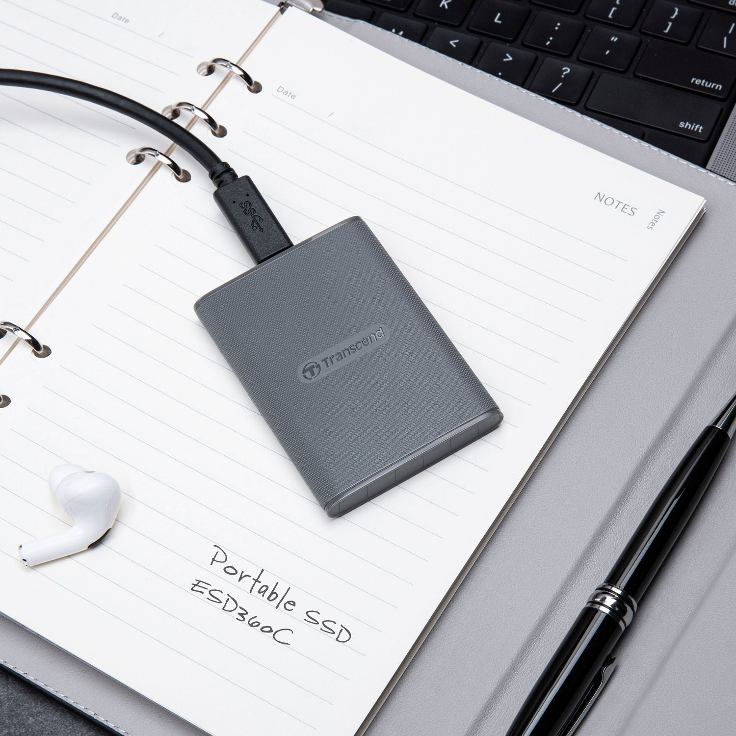 SSD portatile grigio con cavo su taccuino, penna e auricolare nelle vicinanze.