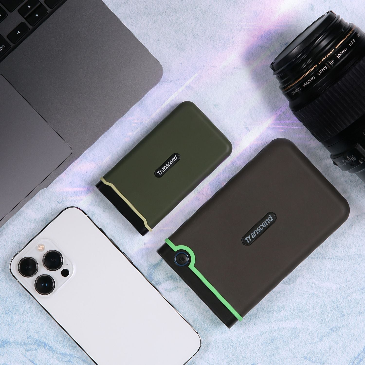 Biały iPhone, dwa zewnętrzne dyski twarde Transcend, laptop i obiektyw Canon.