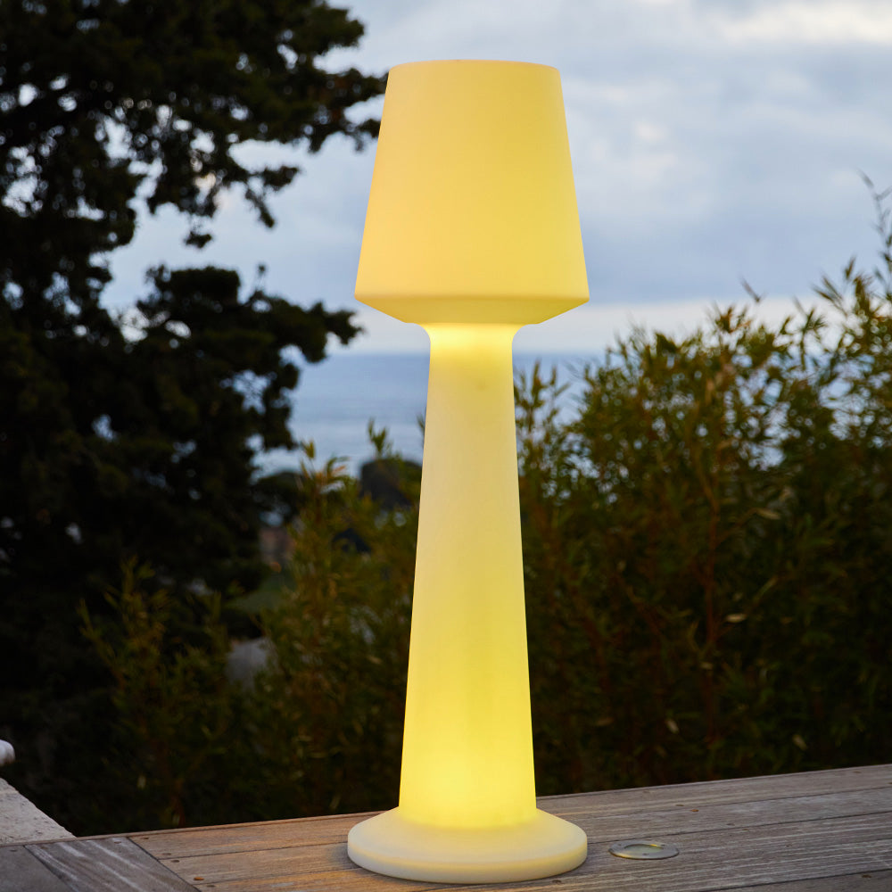 Eine leuchtende Stehlampe steht auf einer Holzterrasse mit Bäumen und Meer im Hintergrund.