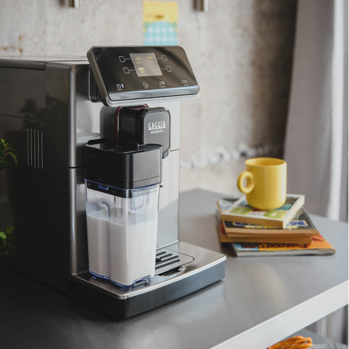 Eine silberne Kaffeemaschine mit Milchtank steht auf einem Tisch, daneben eine gelbe Tasse.
