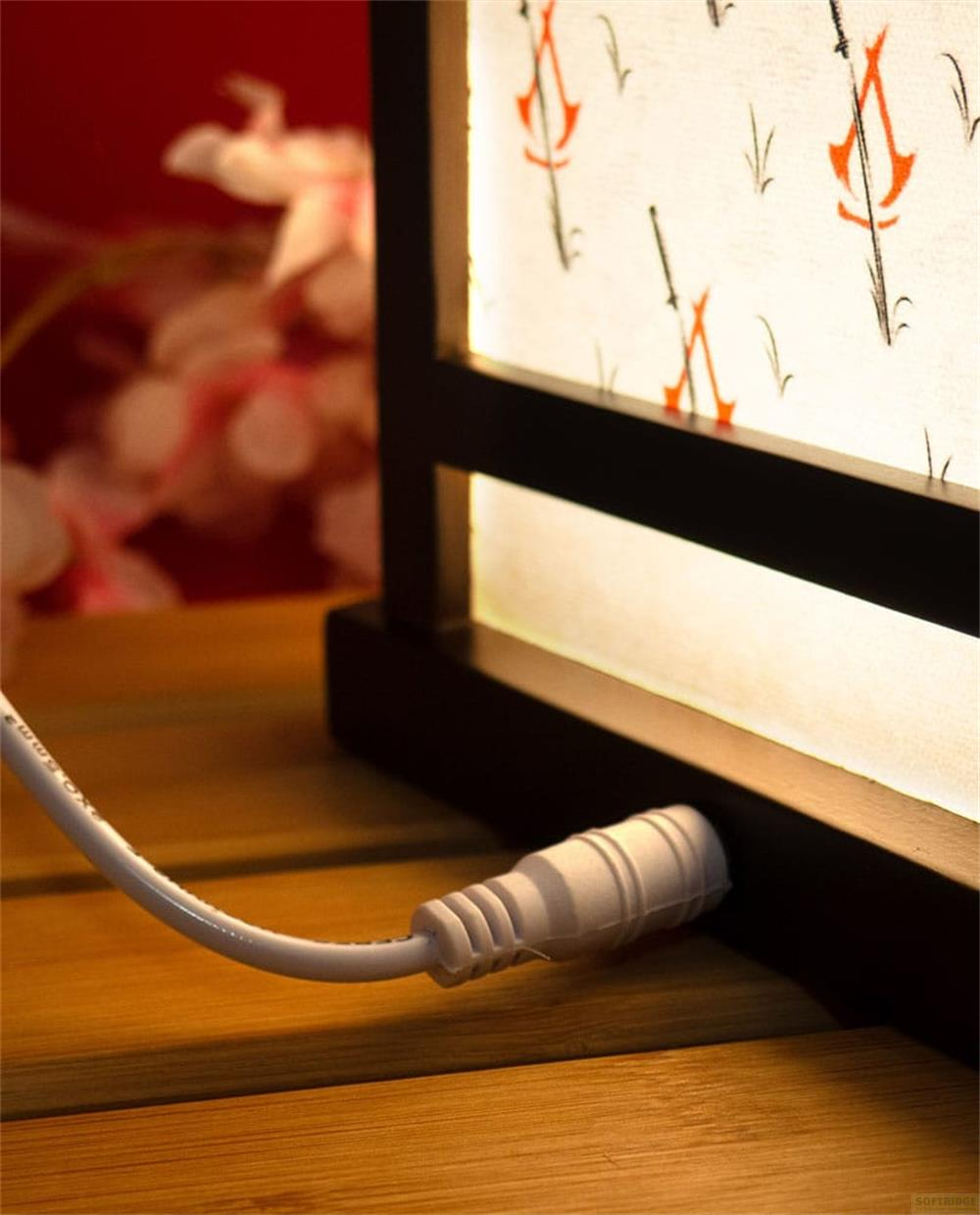 Ein weißes Stromkabel verbindet sich mit einer dekorativen Lampe mit weißem Schirm und orange-schwarzen Mustern, auf einer Holzoberfläche.