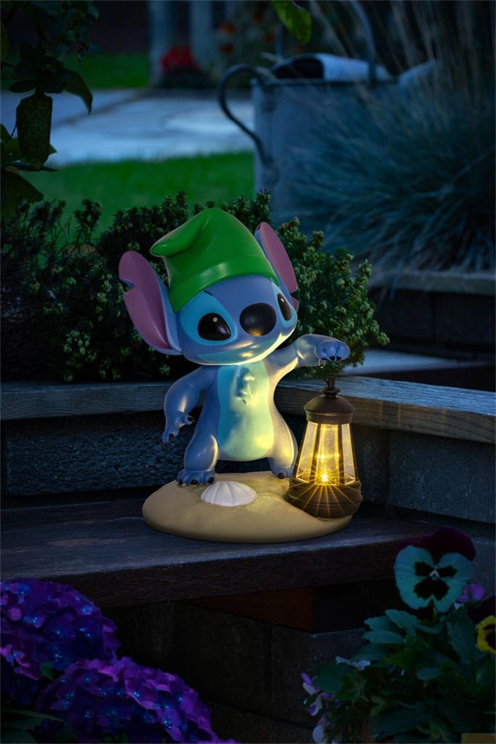 Eine blaue Stitch-Figur mit grünem Hut hält eine leuchtende Laterne im Garten.