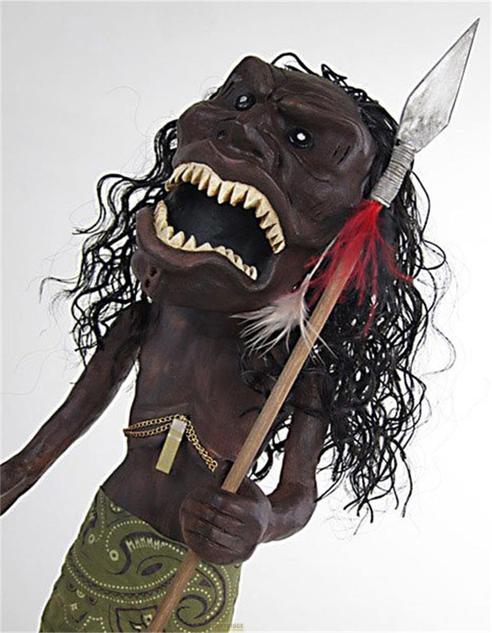 Eine dunkelhäutige, grimmig aussehende Monsterpuppe mit lockigem schwarzem Haar, einem Speer und einem grün gemusterten Rock auf weißem Hintergrund.
