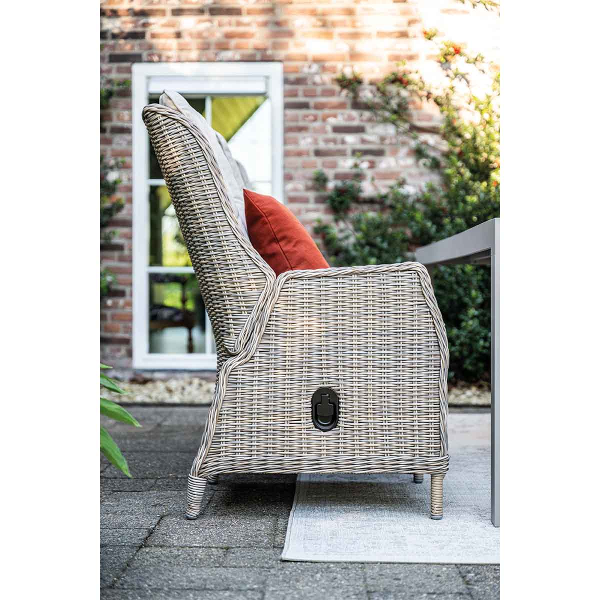 Un fauteuil en rotin gris avec coussin rouge est sur une terrasse en pierre près d'une table.
