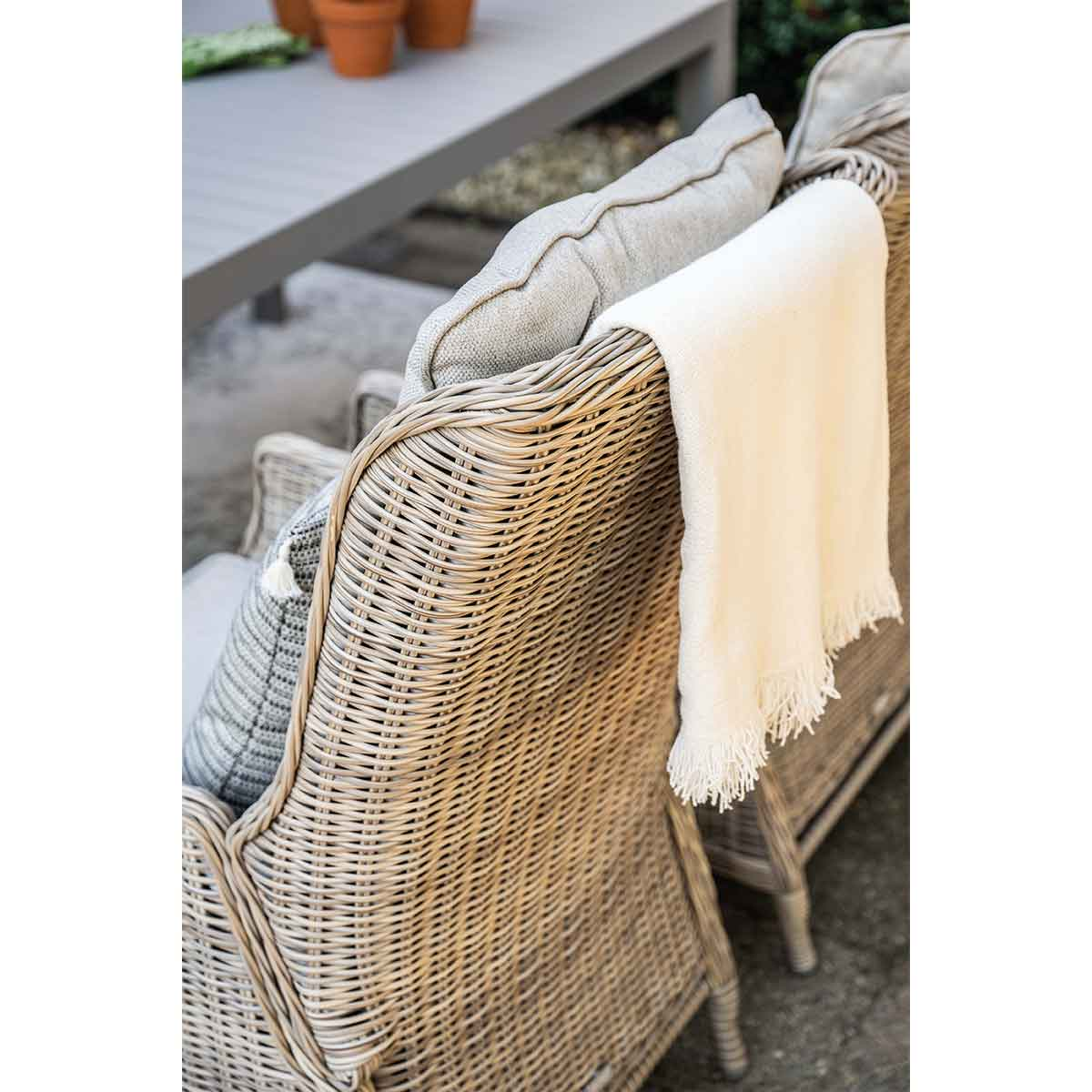 Un fauteuil en osier beige avec coussins et jeté, dehors sur une terrasse.