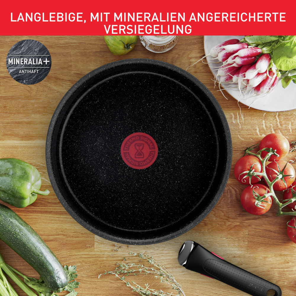 Schwarze Pfanne mit roten Punkten, umgeben von Gemüse und Kräutern.