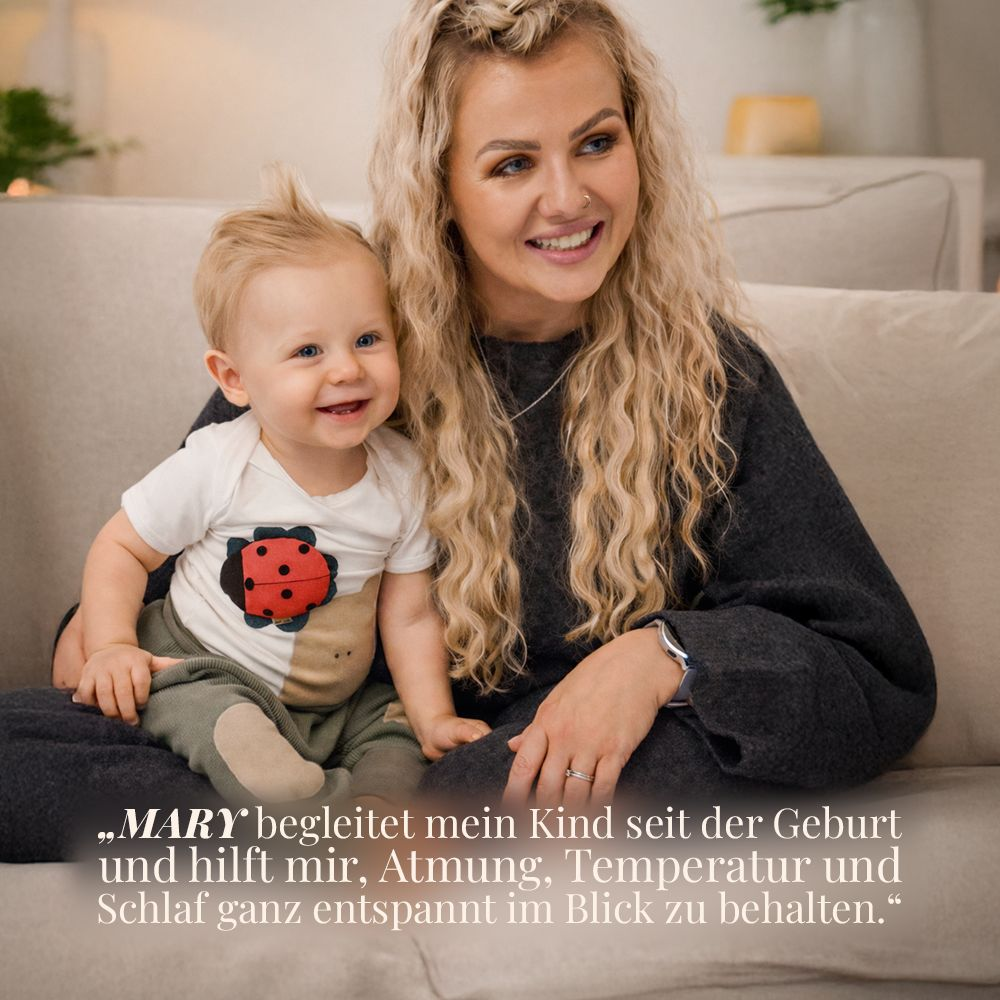 Eine Frau mit blonden Haaren hält ein Baby auf einer Couch. Beide lächeln.
