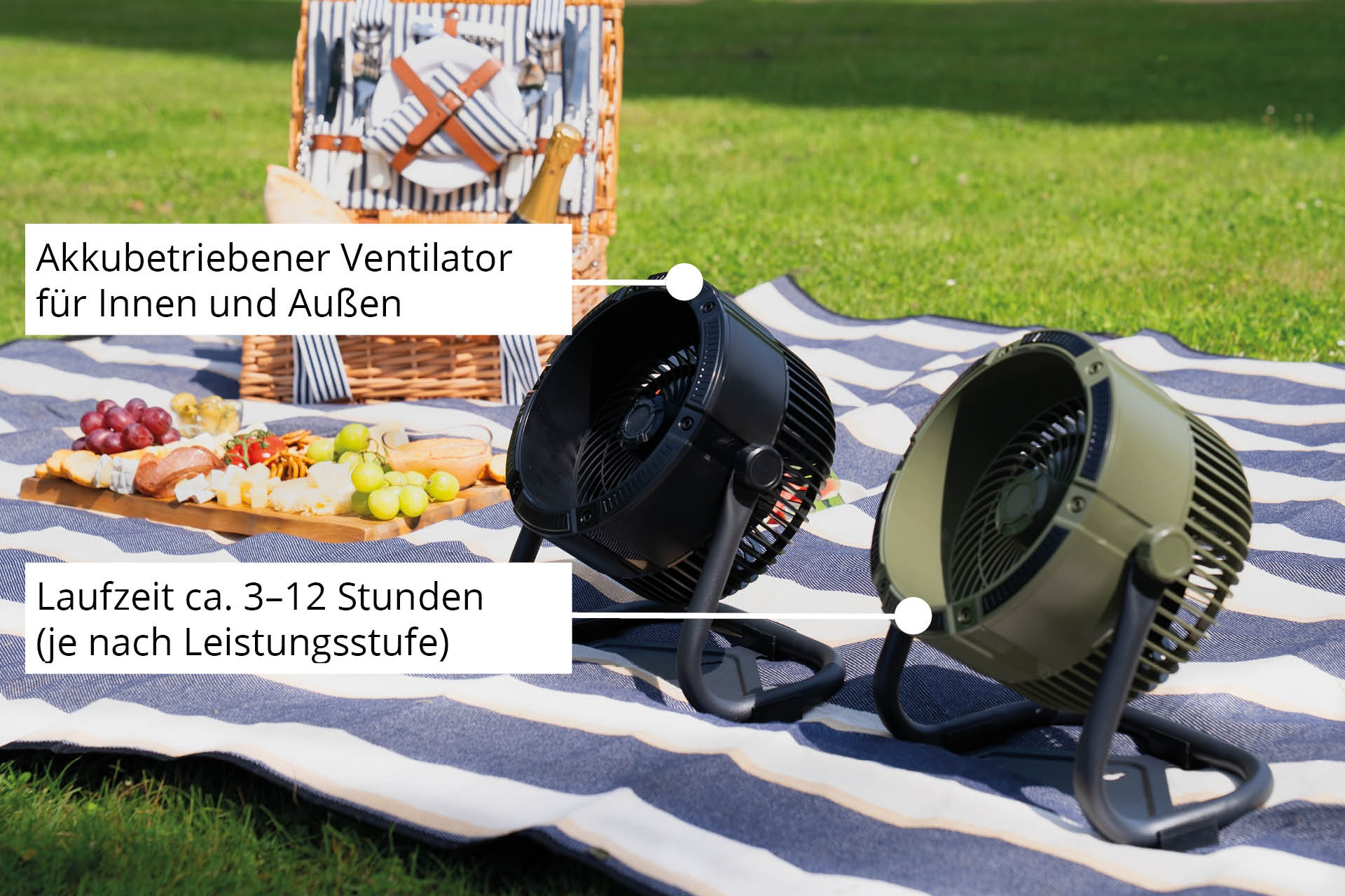 Dwa wentylatory na kocu piknikowym z jedzeniem i koszem piknikowym na trawie.