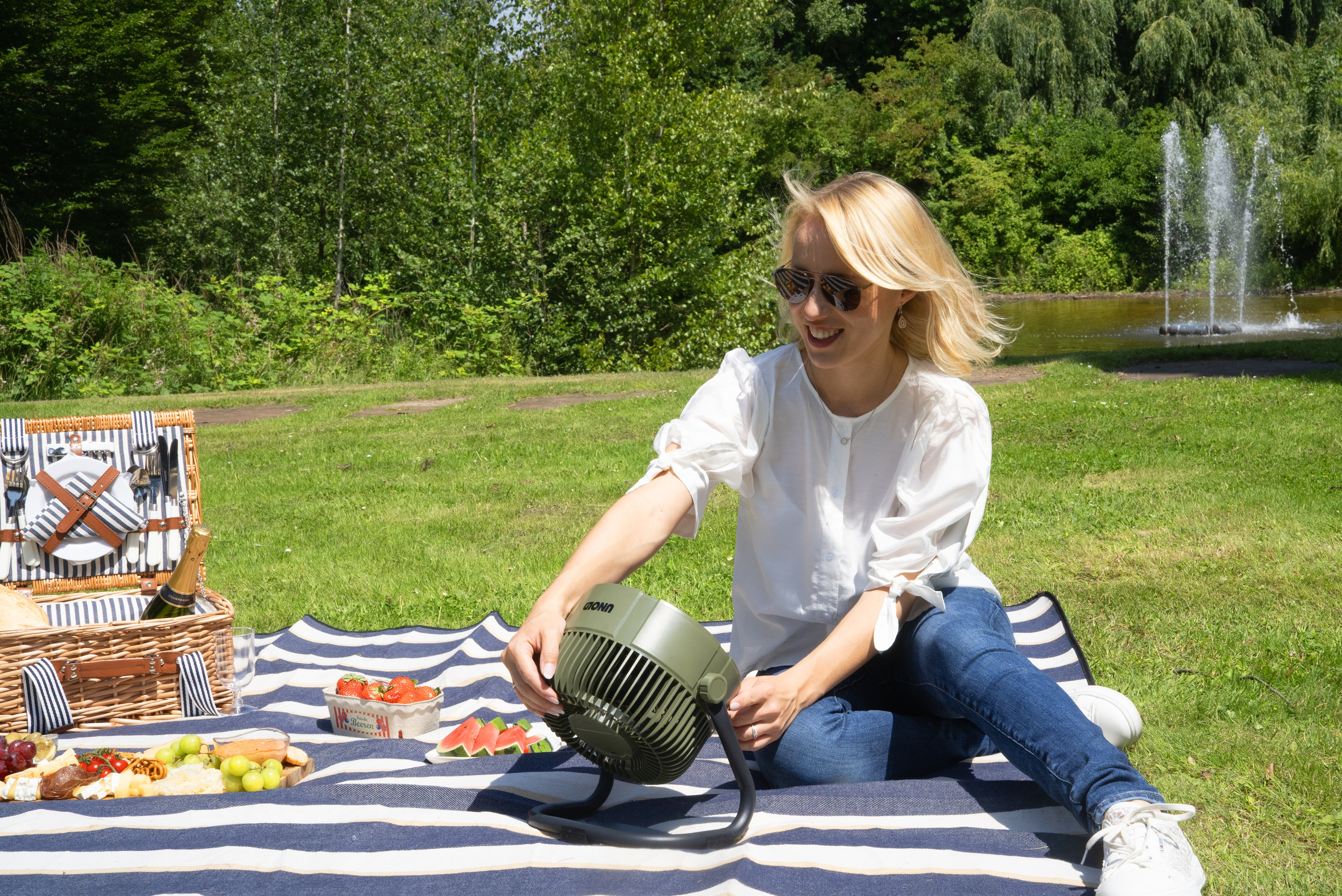 Kobieta z wentylatorem na kocu piknikowym w parku, fontanna w tle.