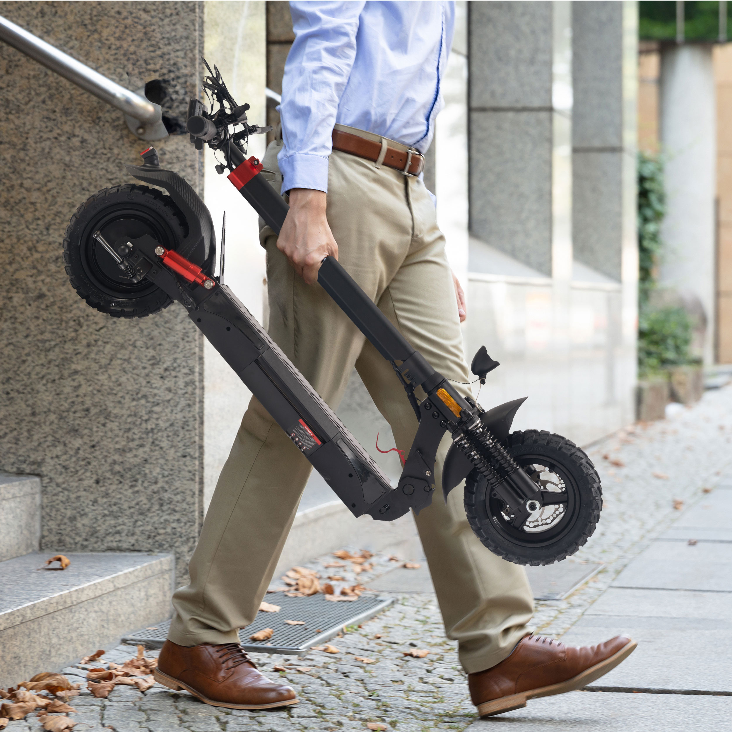 Mann trägt zusammengeklappten Roller, beigefarbene Hose, hellblaues Hemd, braune Schuhe.