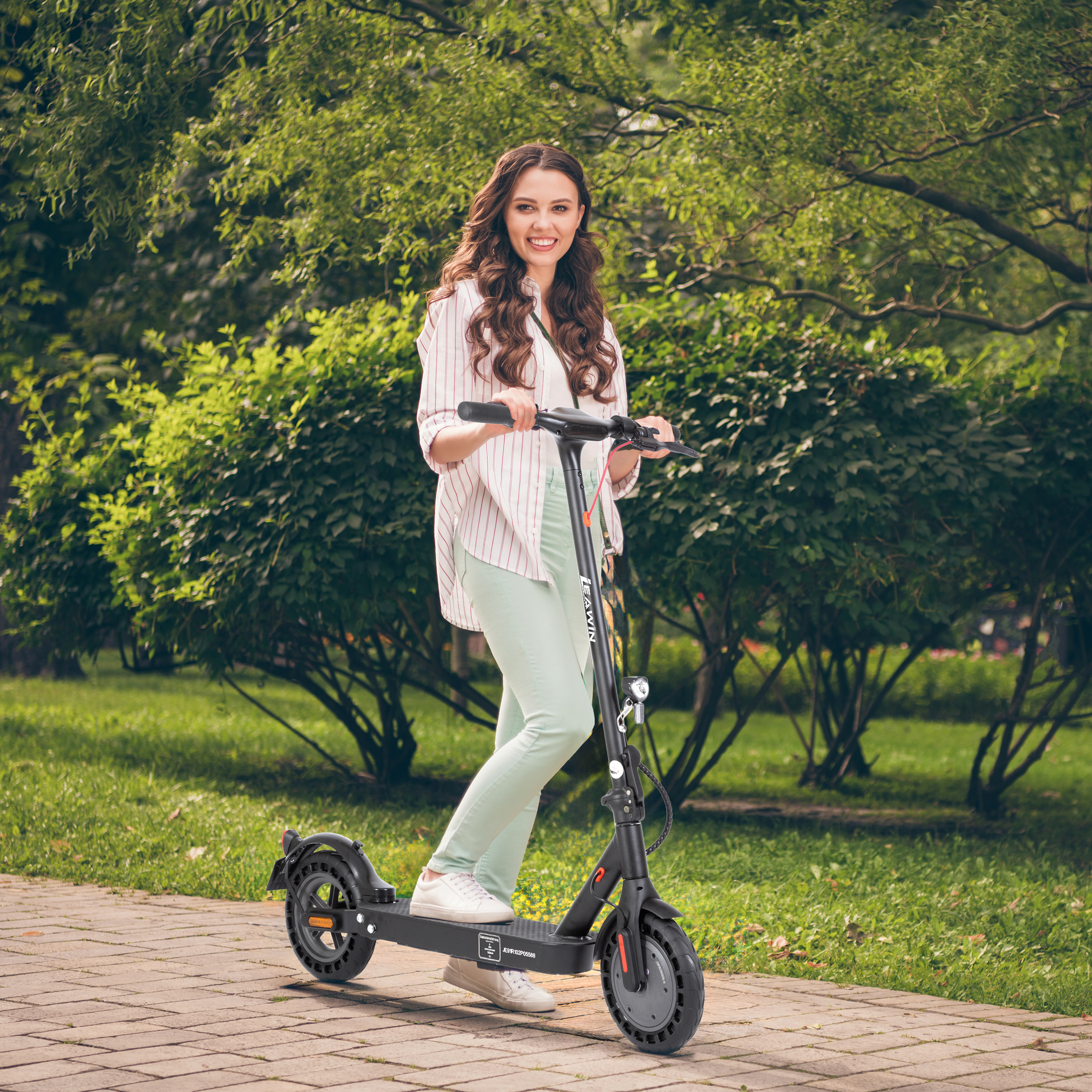 Eine Frau mit langen Locken fährt einen schwarzen Elektroroller auf einem gepflasterten Weg im Park.