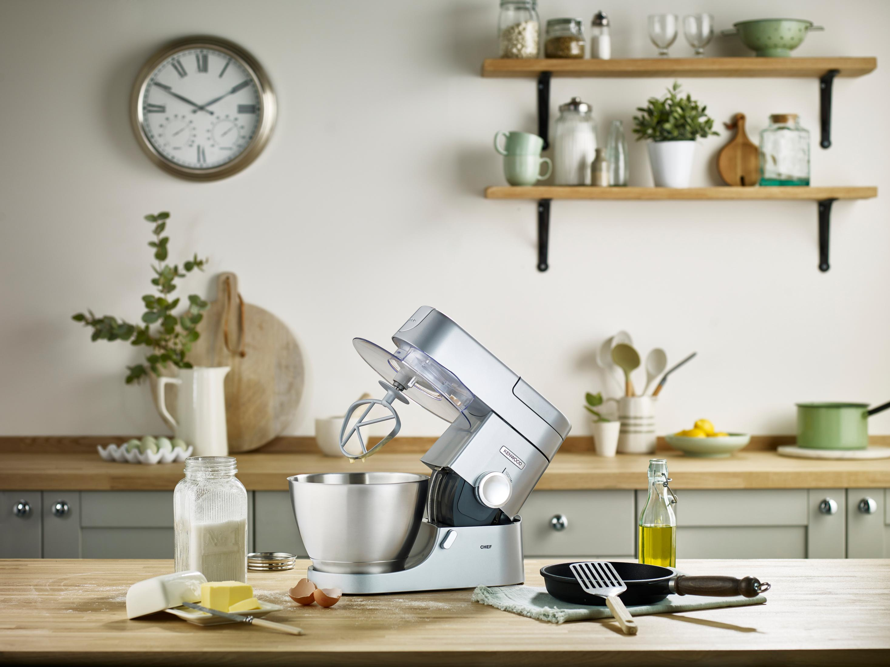 Srebrny robot kuchenny Kenwood stoi na drewnianym stole z takimi składnikami jak masło, jajka i mąka.