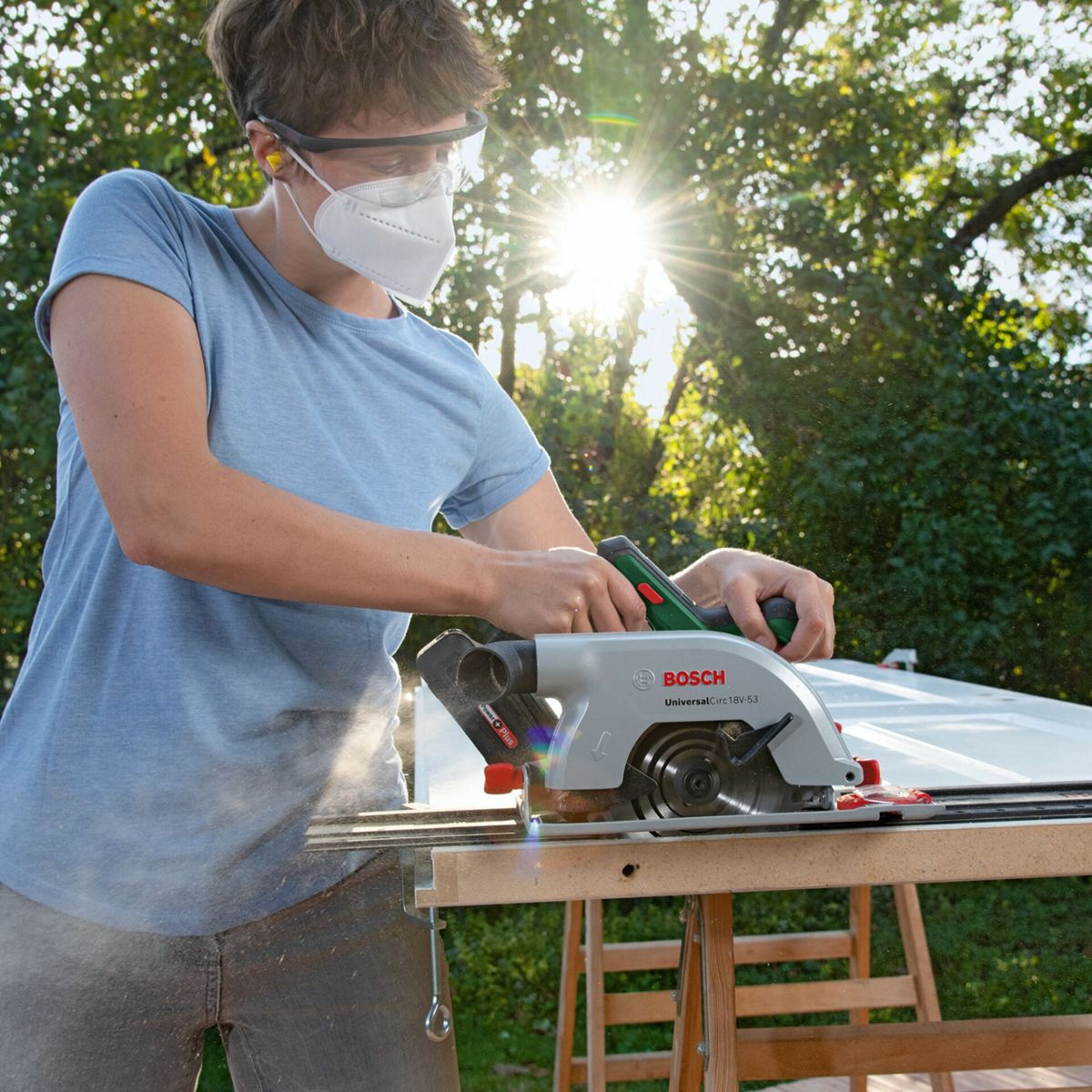 Osoba tnie piłą tarczową na stole na zewnątrz, słońce prześwituje przez drzewa.