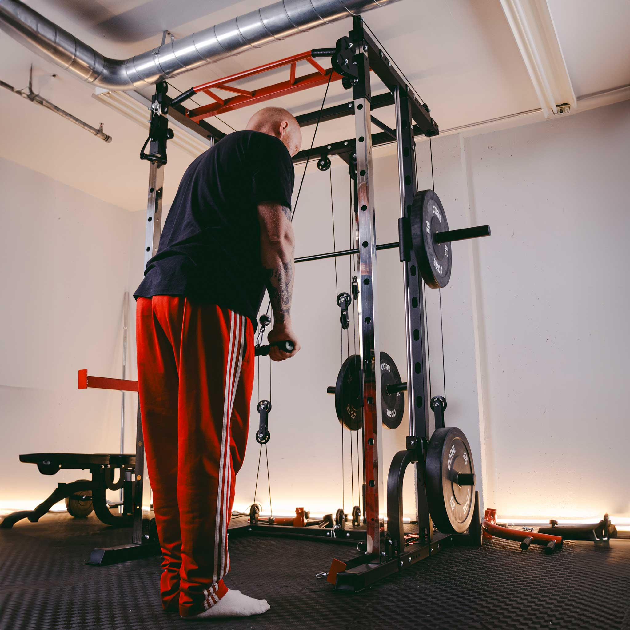 Mężczyzna ćwiczy na maszynie z linkami w siłowni. Nosi czerwone spodnie.