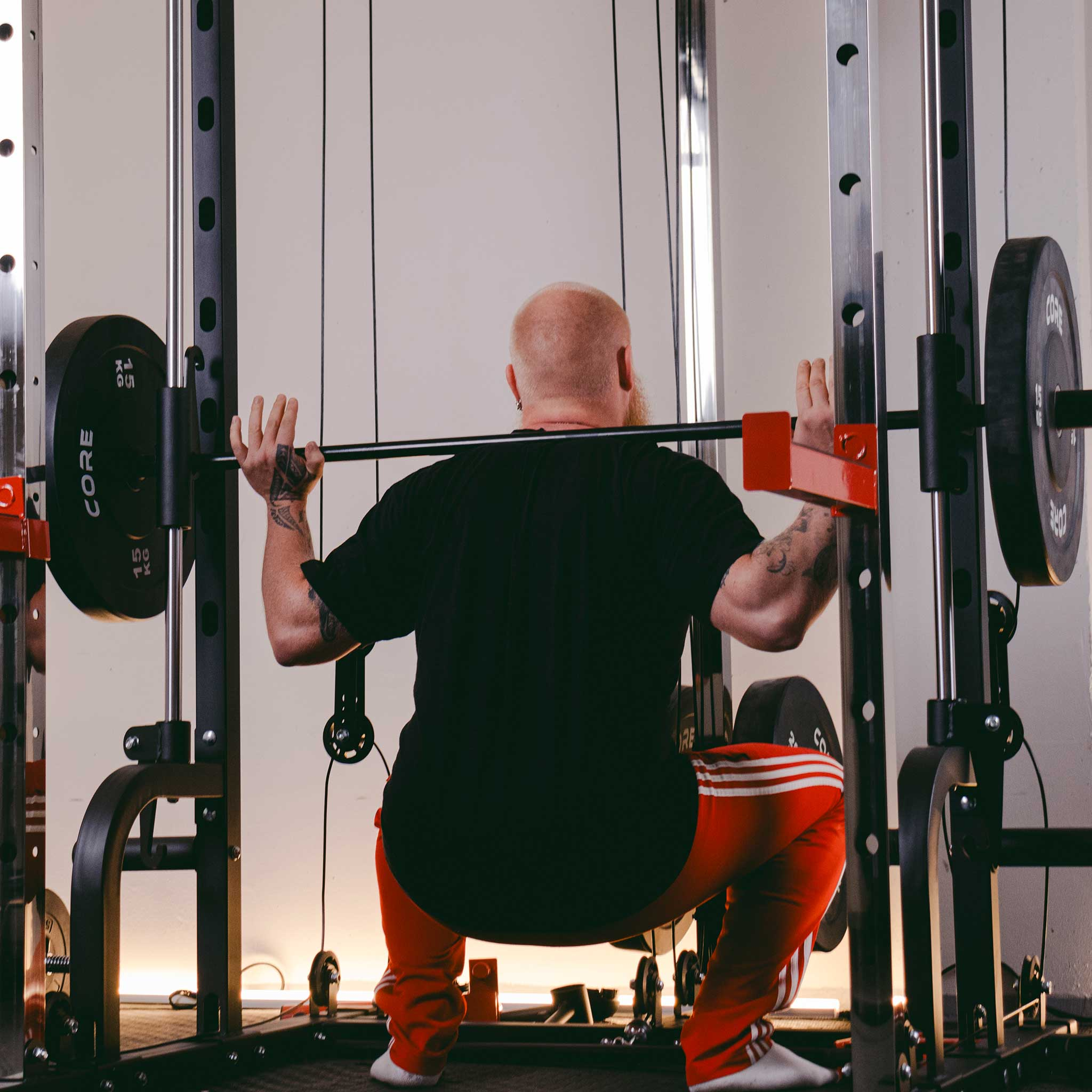 Mężczyzna w czarnej koszulce i czerwonych spodniach robi przysiad ze sztangą.