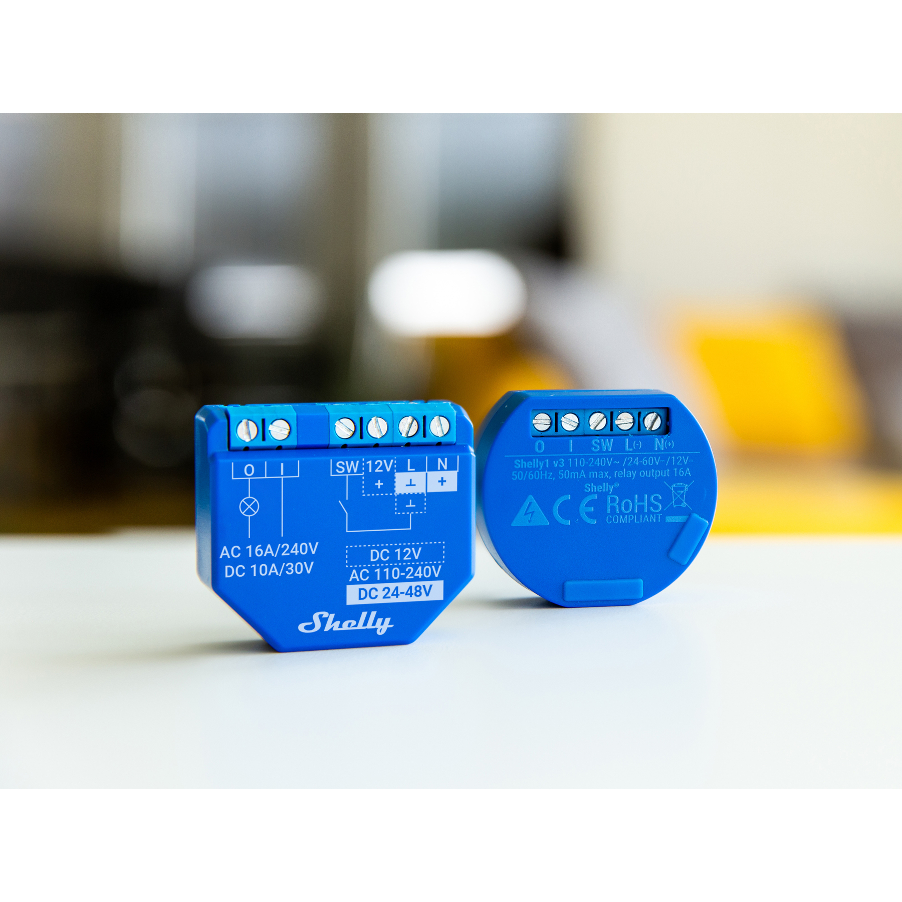 Se muestran dos relés Shelly azules de hogar inteligente, con sus diagramas de cableado y especificaciones técnicas en una superficie blanca.