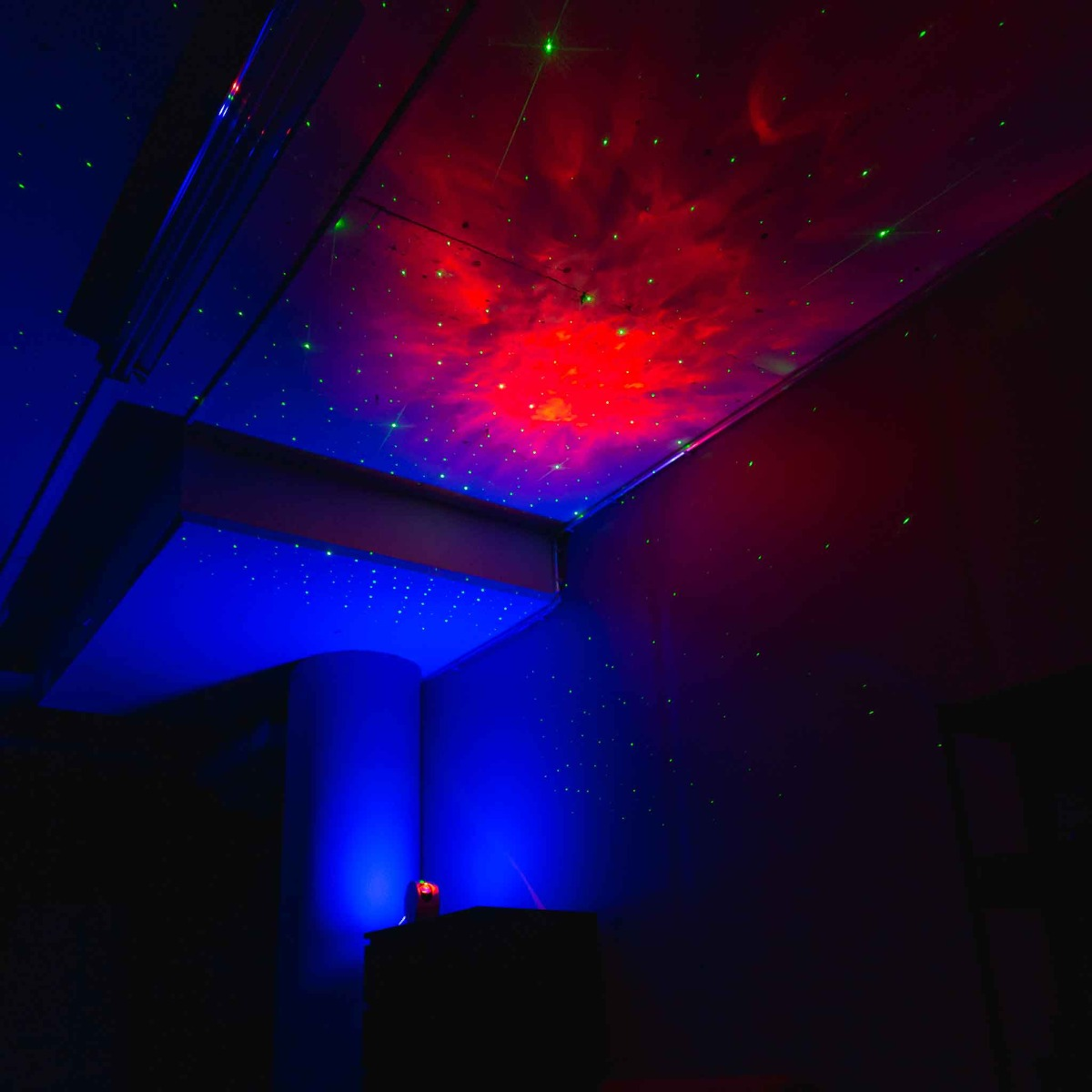 Ein dunkler Raum, beleuchtet von roten und blauen Lichtern, die einen kosmischen Effekt mit grünen Laserpunkten an Decke und Wänden erzeugen.