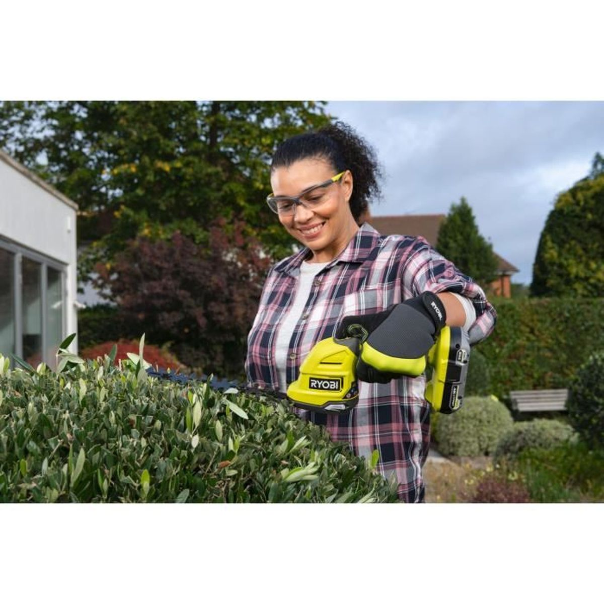 Frau schneidet Hecke mit gelber Ryobi-Schere, trägt Brille und kariertes Hemd.