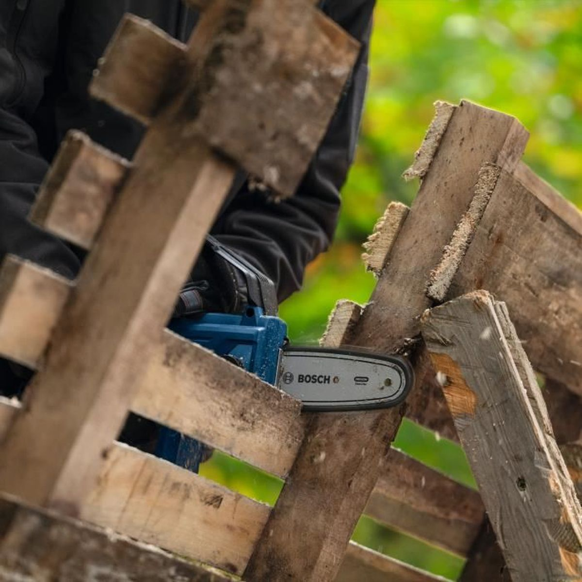 Eine Person benutzt eine blaue Bosch-Kettensäge, um Holzplanken im Freien zu schneiden.