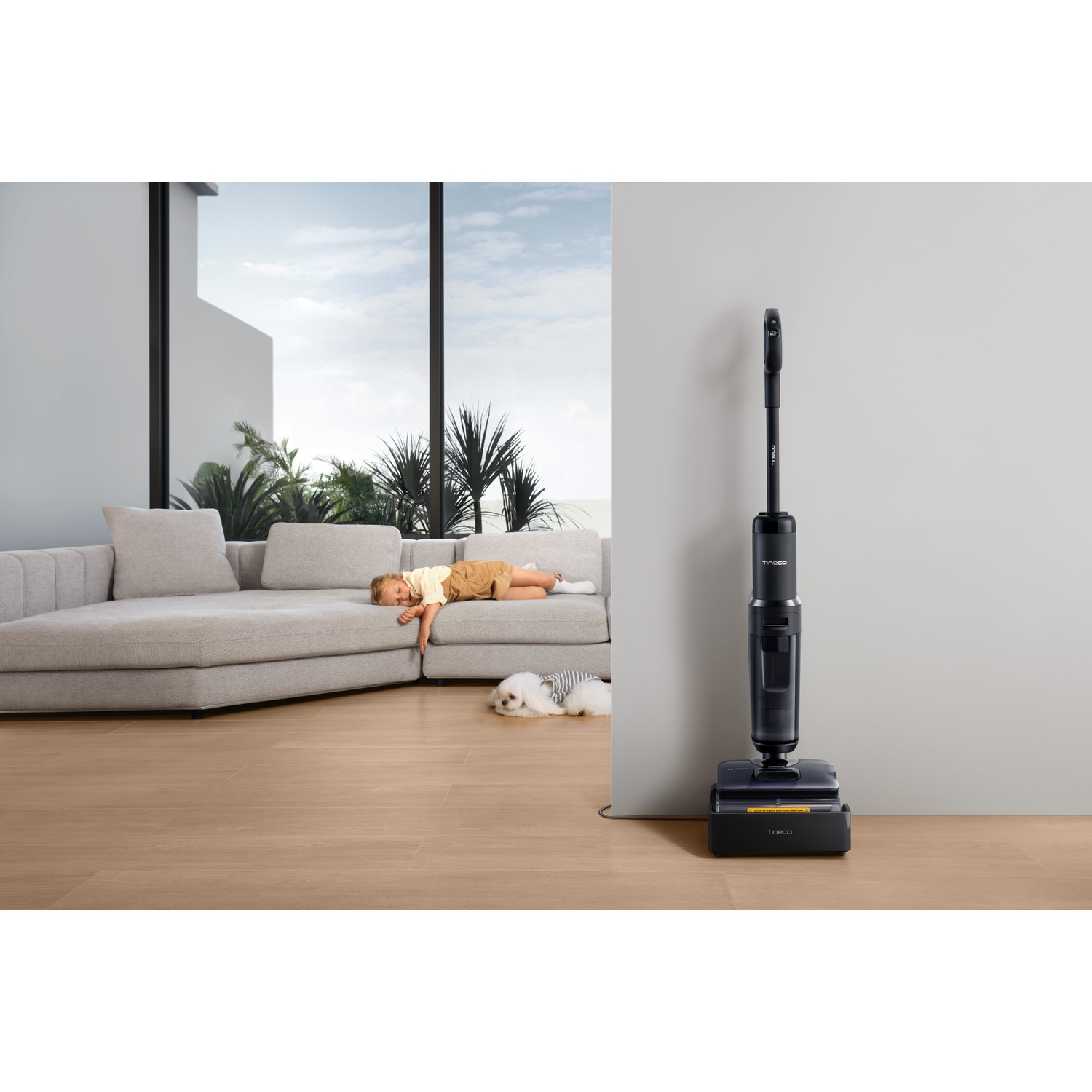 Ciemny odkurzacz stoi na stacji ładującej obok sofy, na której odpoczywa dziecko i pies.