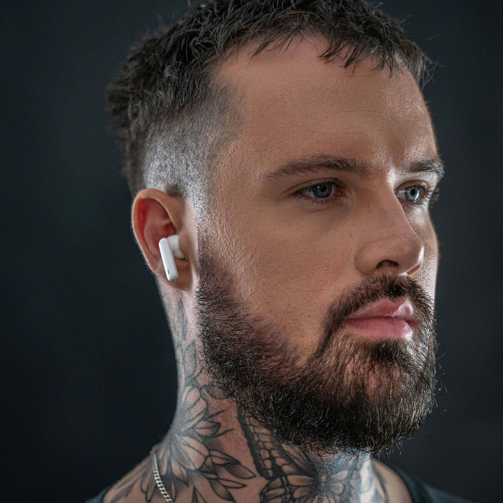Mężczyzna z brodą i tatuażami nosi białe słuchawki.