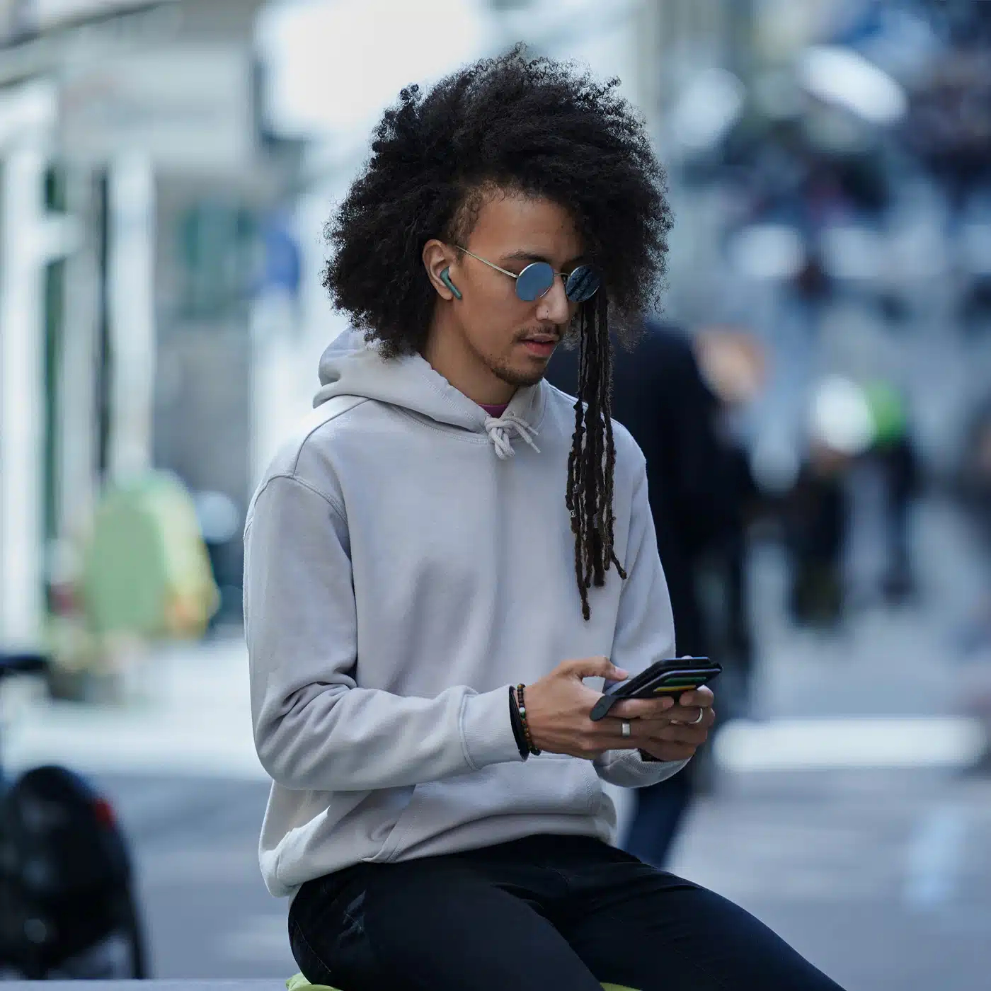 Mann mit Afro und Dreadlocks trägt Sonnenbrille und Kapuzenpullover, benutzt Handy.