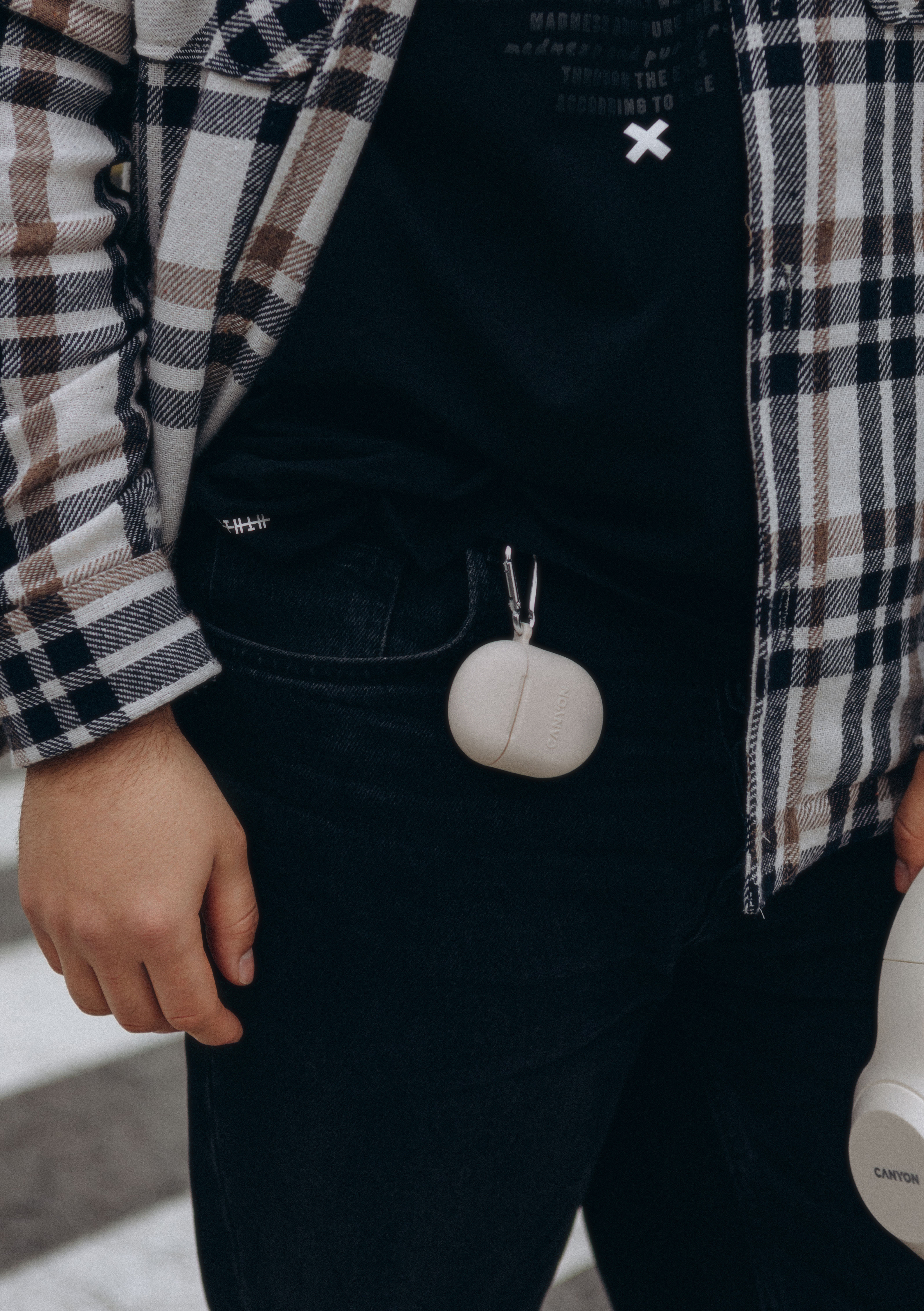 Osoba w kraciastej koszuli i ciemnych dżinsach, z beżowym etui na słuchawki przypiętym do szlufki.