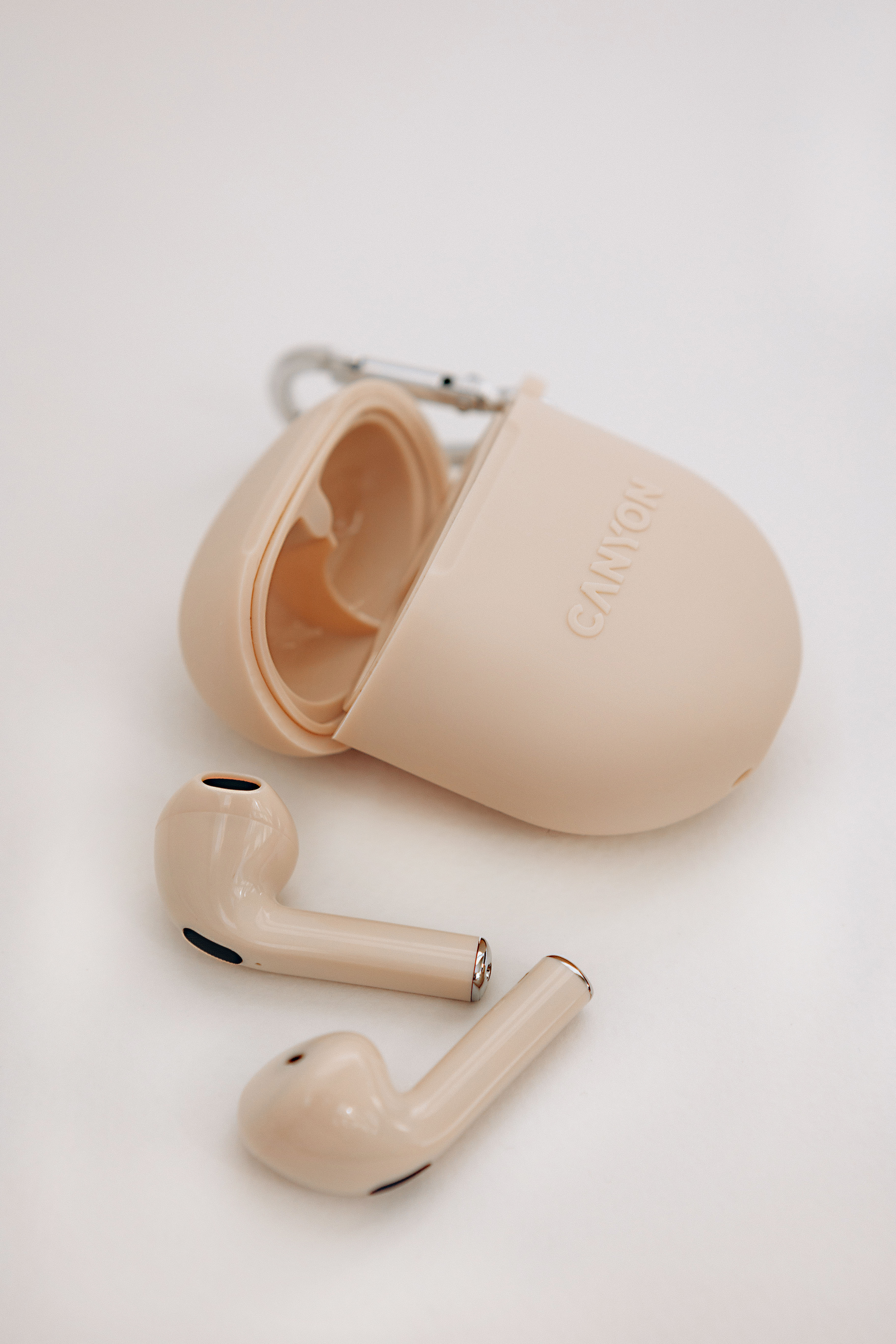 Beżowe słuchawki obok otwartego etui ładującego z logo Canyon na białym tle.